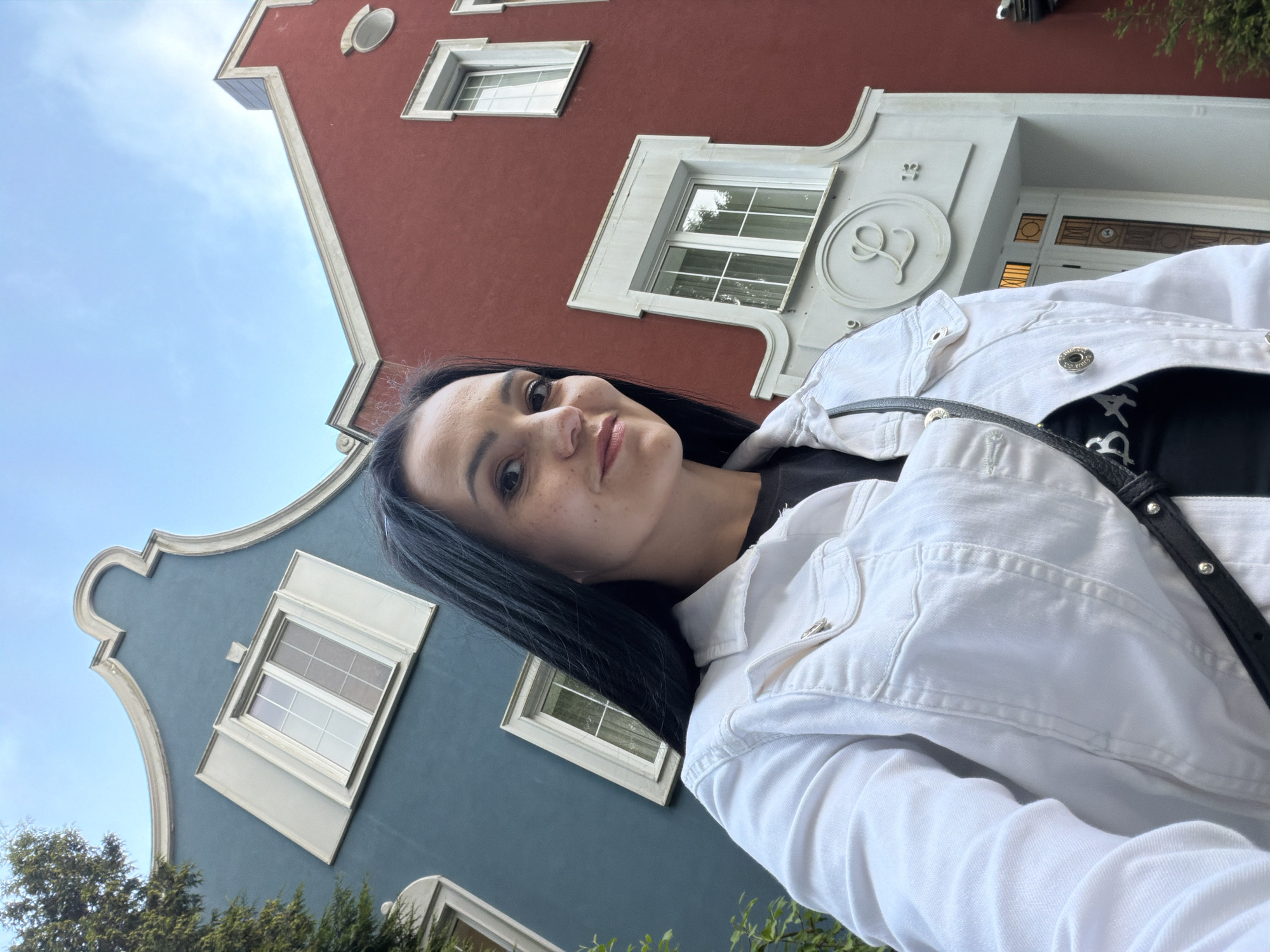 A woman with long black hair and fair skin, wearing a white denim jacket over a black t-shirt, stands in front of two buildings. The building on the left is painted teal and features a curved gable with a white trim and a single window. The building on the right is painted red and has a more angular roofline, also with white trim, and multiple windows. The woman is looking towards the camera, with her face turned slightly to her right. She is wearing makeup and has freckles on her nose and cheeks. A black bag with silver studs is slung across her body. The sky above is a light blue with a few wispy clouds. The architectural style of the buildings, particularly the gables and decorative elements, suggests they are located in Svetlogorsk, Russia, a resort town known for its distinctive architecture.