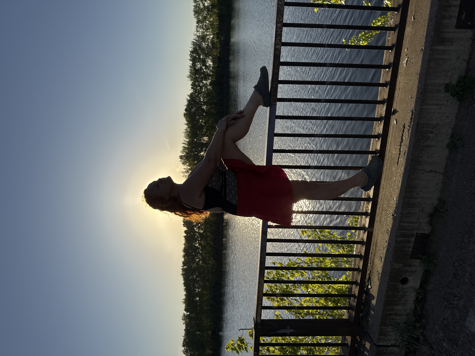A person is standing on a bridge with one leg outstretched and resting on the bridge's railing. The other leg is bent and the foot is held in the person's hand. They are wearing a dark top and a short red skirt. The bridge has a metal railing with thin vertical bars. Behind the bridge is a body of water, and beyond that are trees. The sun is behind the person, creating a silhouette effect. The location may be Chaikovsky, Russia.