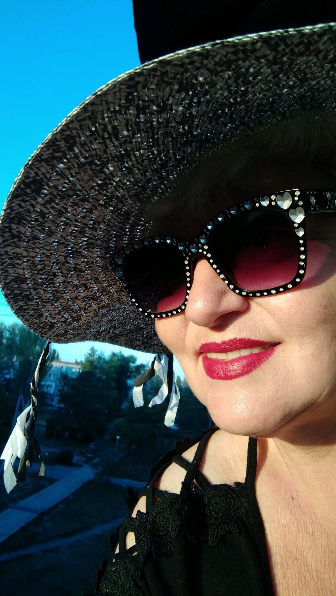 A woman is posing for a selfie outdoors in front of a blue sky and a blurred background of trees and buildings. She is wearing a large, patterned sun hat adorned with rhinestones, and oversized sunglasses that are also decorated with rhinestones in a heart shape. Her lips are painted with bright red lipstick. She is wearing a black top with intricate lace detailing on the shoulders. The image appears to be taken in the city of Horishni Plavni, Ukraine, possibly from a high vantage point overlooking the city.