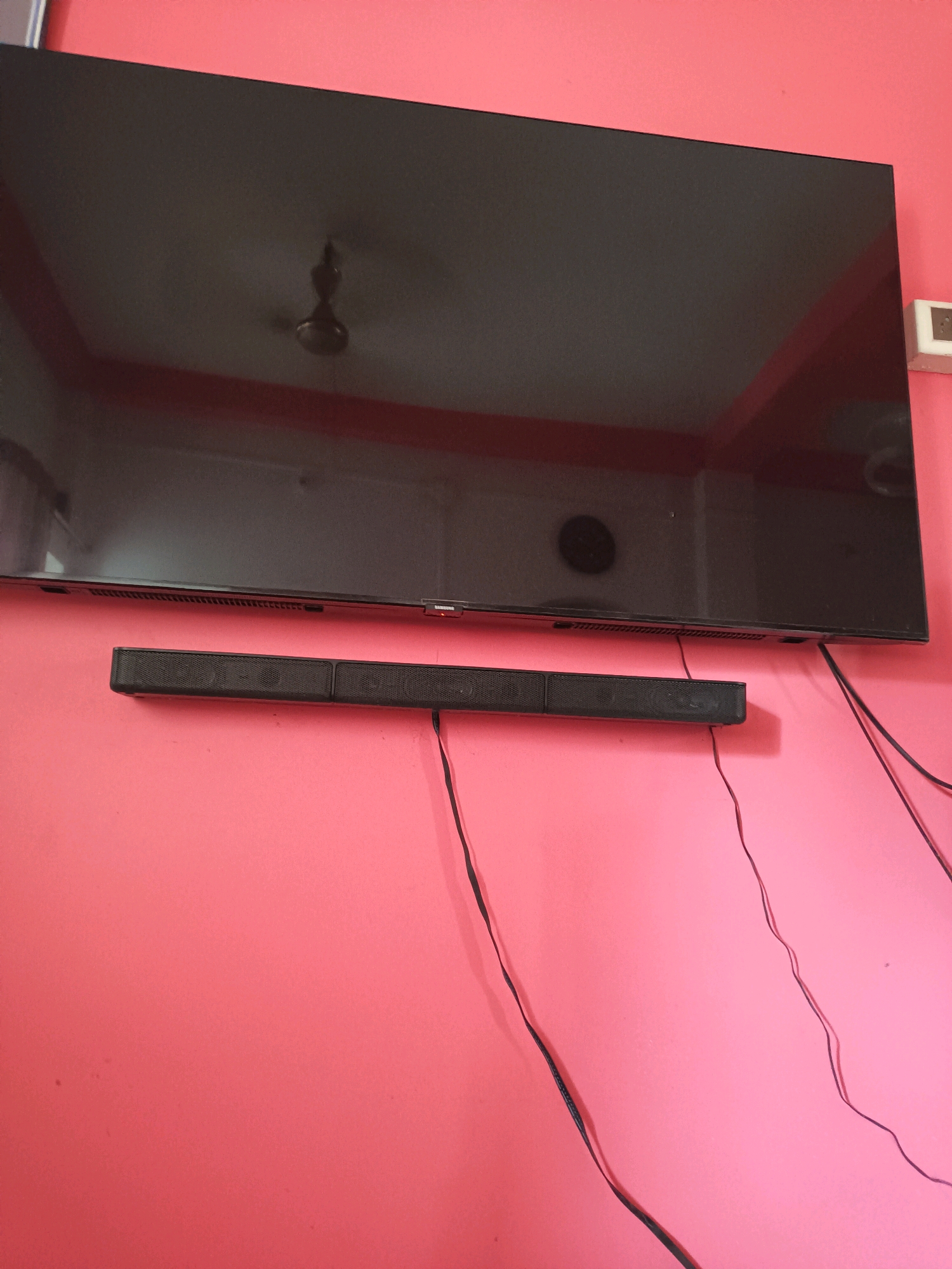 A wide, flat-screen television is mounted on a bright pink wall. Below the television, a black soundbar is also mounted on the wall. Several black wires are visible, some connected to the soundbar and others hanging loosely. A ceiling fan is reflected in the dark screen of the television. The image appears to have been taken indoors, likely in a residential setting. There is no information about the location in Gaighata, India, or any people.