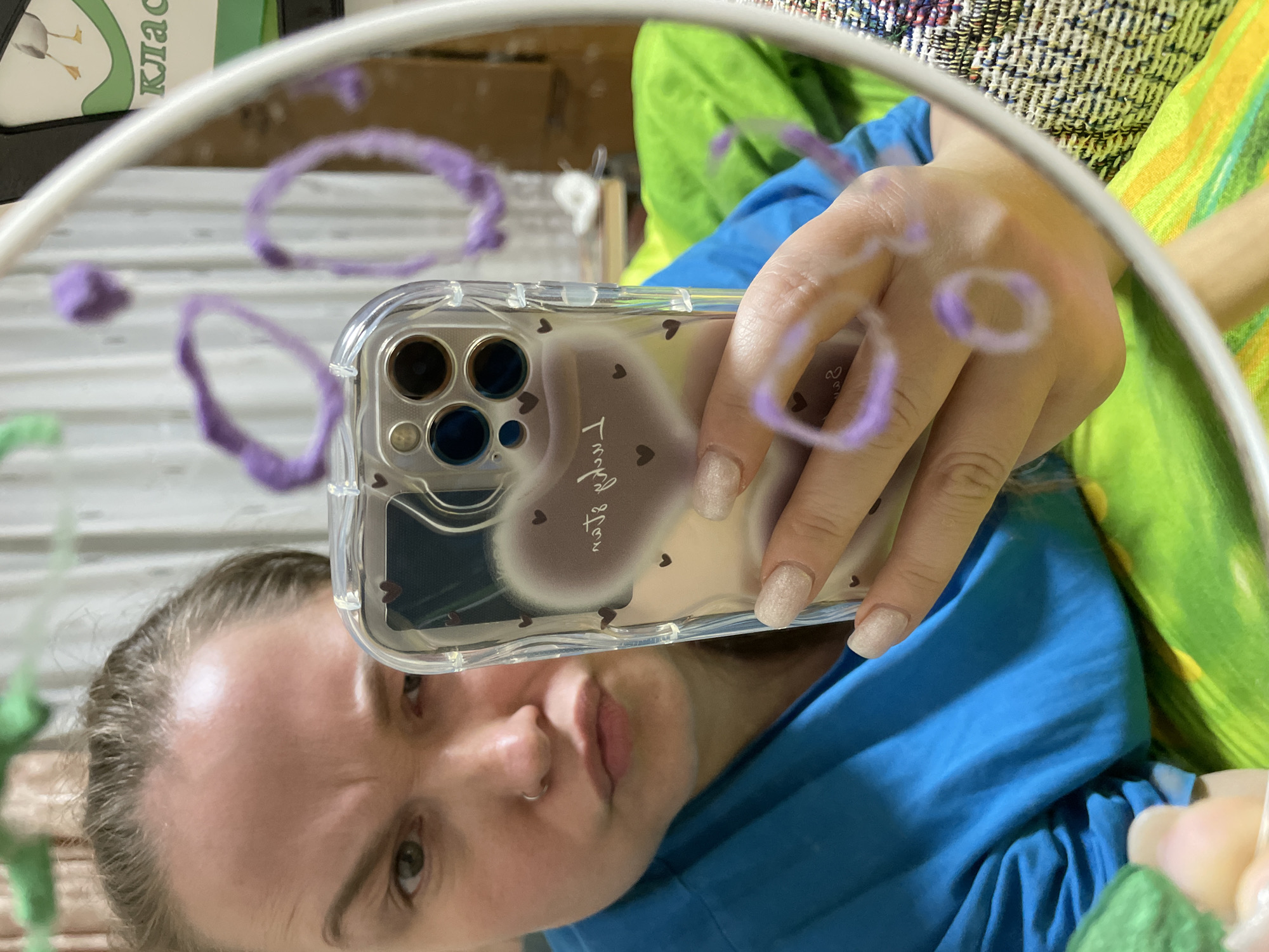 The media file captures a mirror selfie, presenting an inverted reflection of a person holding a smartphone. The subject, a person with light skin and light brown hair, wears a blue top, possibly a hoodie, and has a small nose ring. Their long fingernails are painted with a light, possibly glittery, polish. The person's expression is neutral, looking directly at the phone's camera.
The smartphone, identifiable by its multiple camera lenses, is encased in a clear cover decorated with small dark heart shapes. Faint, stylized cursive text, appearing as "Thank you" or similar, is subtly visible on the phone case through the reflection. A blurry, light-colored, fluffy object is reflected on the phone's screen.
Several loops and strands of purple yarn or thread are either attached to or drawn on the mirror's surface, appearing in the foreground and interacting with the reflected image. The mirror itself has a thin white frame.
The background, viewed through the mirror, includes a vibrant green and yellow patterned fabric, likely a cushion or blanket, and another textured woven fabric. In the upper left corner of the reflection, a white object with a green top displays partially visible Cyrillic text, "Клас" (Klass), which means "Class."
The scene is set indoors, under ambient daylight, likely in a personal space such as a bedroom or living area in Izhevsk, Russia. The image captures a candid moment of self-portraiture with playful decorative elements on the mirror.
