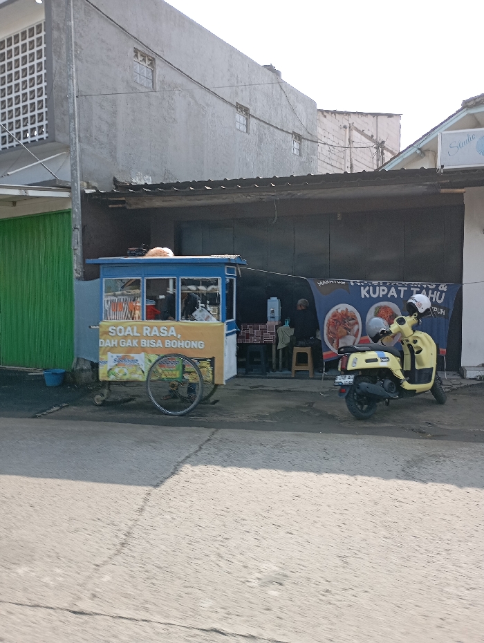 A daytime street scene in Pataruman, Indonesia, depicts a commercial food vendor setup and a parked motorcycle. In the mid-ground, a blue and yellow mobile food cart, bearing the Indonesian text "SOAL RASA, DAH GAK BISA BOHONG" (Regarding taste, it cannot lie), is stationary on an unpaved shoulder beside a grey asphalt road. A blue bucket rests near the cart's left side, adjacent to a closed green corrugated metal shutter.
Directly behind the food cart, an open-front food stall operates under a dark corrugated metal roof. Two individuals are present within this stall; one is seated on a stool at a table, and another is partially obscured behind a counter. A large, partially visible banner, predominantly blue and white, displays the words "KUPAT TAHU" and images of food items.
To the right of the food stall, a bright yellow scooter-style motorcycle is parked on its kickstand, with two white helmets resting on its seat.
The background features a multi-story building with a grey, textured exterior and multiple small, gridded windows. Utility lines are visible spanning across the upper section of the image. A partial view of an adjacent building with a white facade and a sign reading "Studio" is visible on the far right. The scene is illuminated by direct sunlight, casting distinct shadows on the road surface in the foreground.