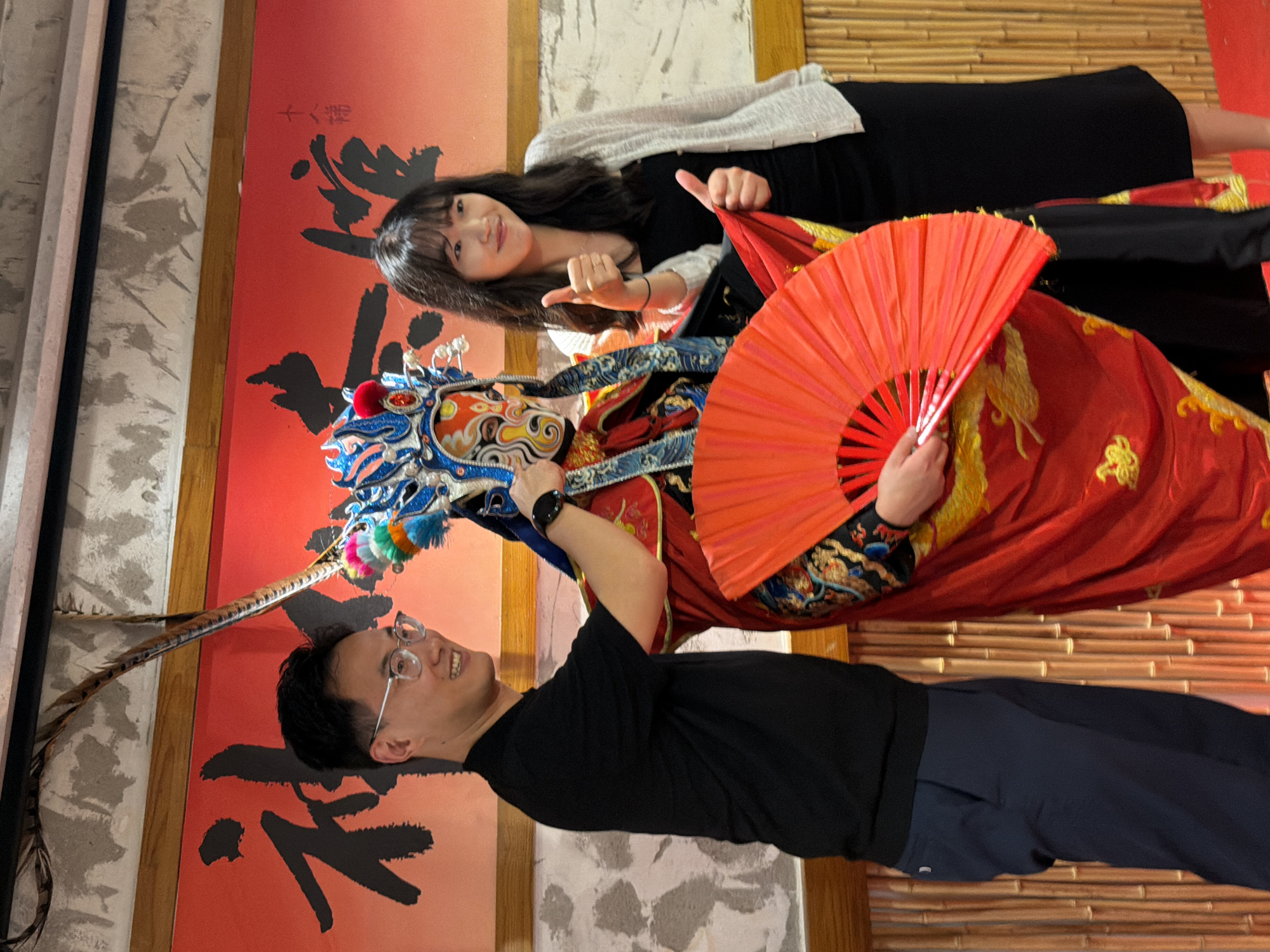 In Yuzhong District, China, three individuals pose in front of a red backdrop with black Chinese calligraphy. On the left, a man wearing glasses, a black t-shirt, and dark blue trousers smiles at the camera, with his right arm extended towards the center. In the middle, a performer in elaborate traditional Chinese opera attire, including a colorful mask and headdress with feathers, holds a large red fan with both hands. The performer's face is obscured by the mask, but they are wearing a red and gold costume. On the right, a woman with long dark hair, wearing a white cardigan over a black dress, gives a double thumbs-up gesture. The backdrop is adorned with bamboo-like textures at the bottom and a smooth concrete-like texture at the top. The lighting suggests an indoor setting, possibly a restaurant or cultural venue.