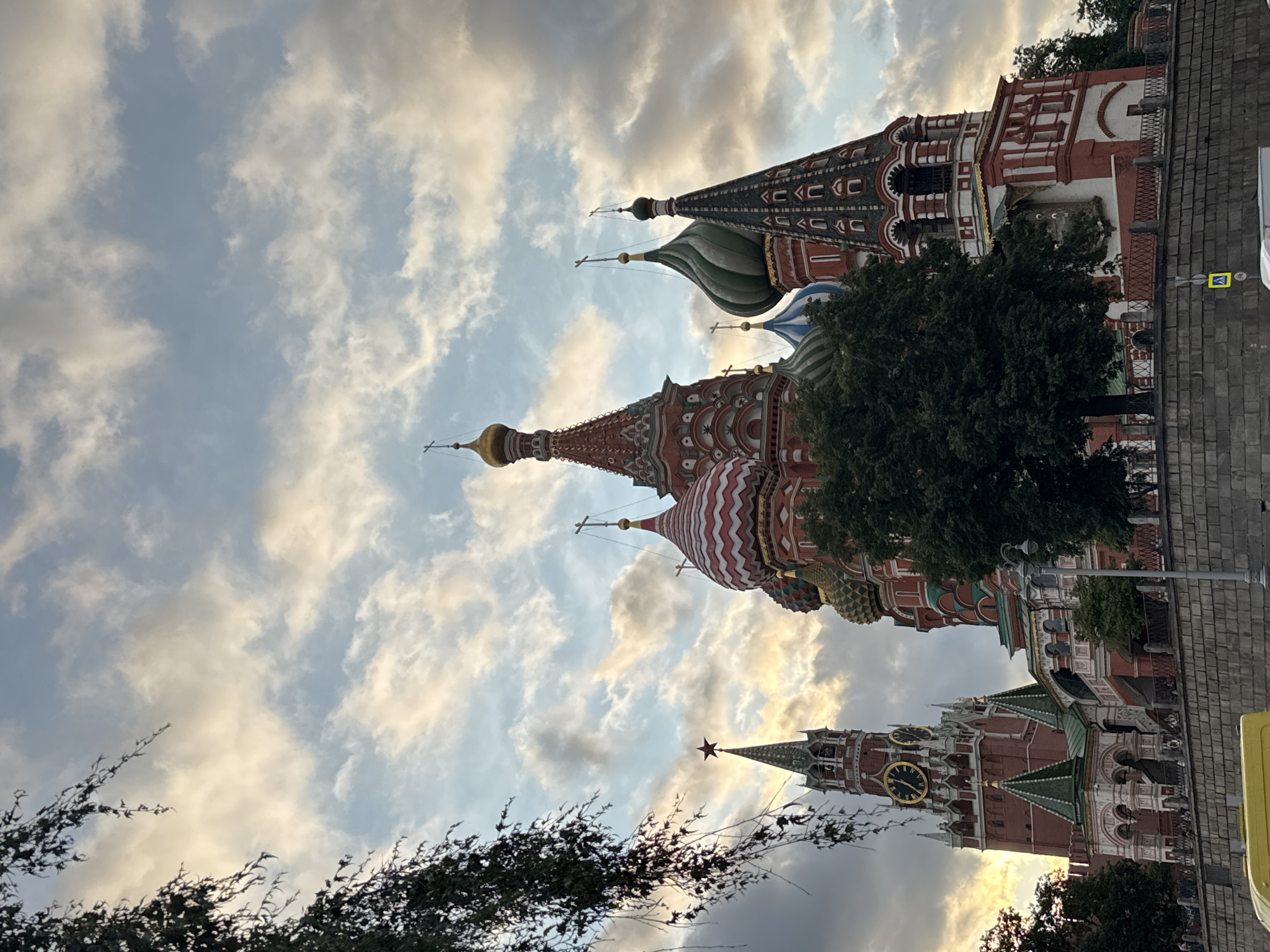 The image captures a low-angle view of St. Basil's Cathedral and the Spasskaya Tower in Moscow, Russia, under a dramatic sky filled with clouds. The iconic onion domes of St. Basil's Cathedral, with their vibrant red, white, and green patterns, are prominently featured. To the left, the Spasskaya Tower, part of the Moscow Kremlin, stands tall with its clock faces and a red star at its peak. A large, dark green tree is situated in the foreground, partially obscuring the view of the cathedral. A stone wall runs along the bottom of the image, with a street and some yellow vehicles visible behind it. The lighting suggests it is either sunrise or sunset, with warm hues illuminating the clouds. No people are clearly discernible in the frame.