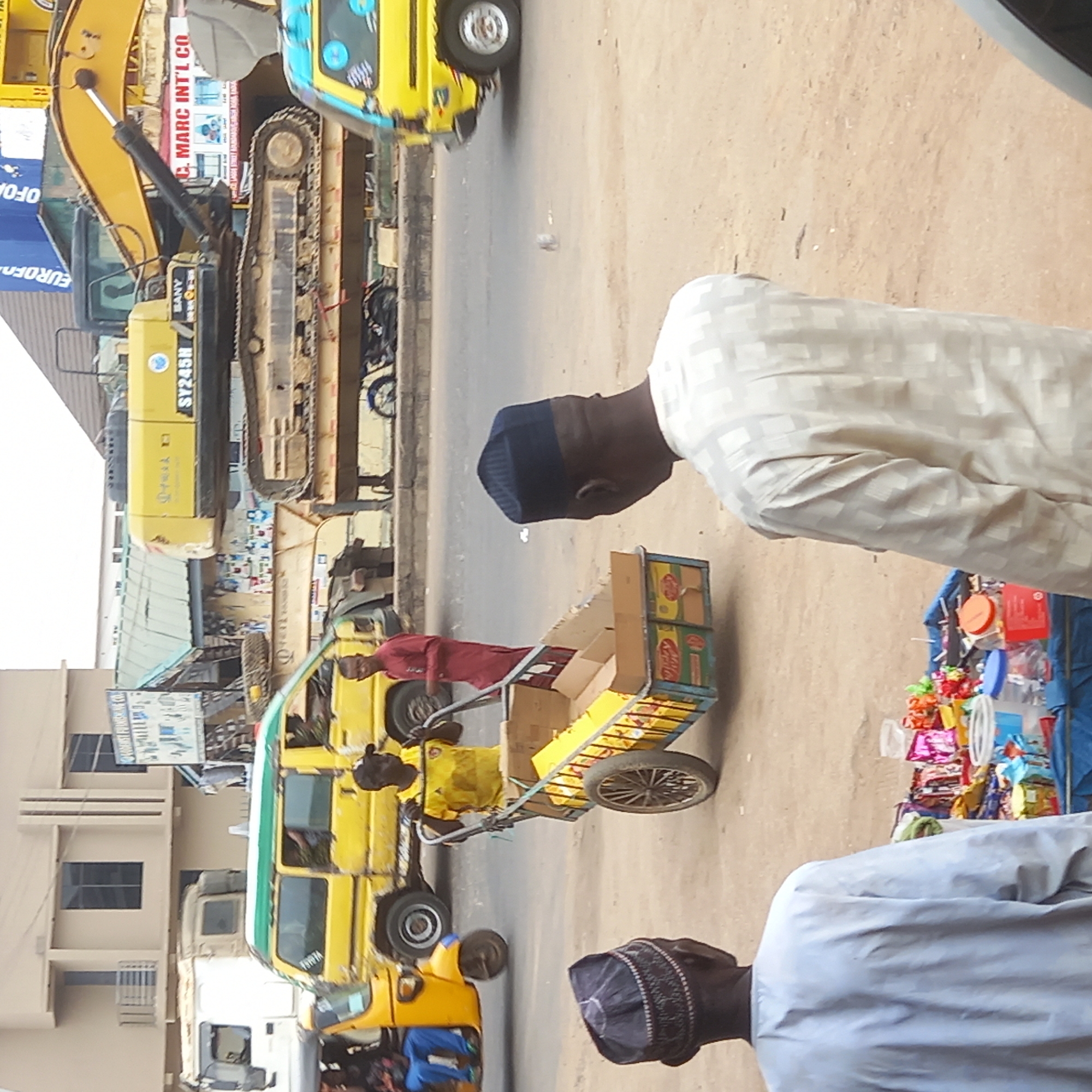 The image depicts a bustling street scene in what appears to be a commercial or industrial area in Dadi Riba, Nigeria. The photograph captures everyday activity under bright daylight conditions.
In the foreground, two men stand facing away from the viewer. One wears a light-colored, geometrically patterned long-sleeved shirt and a dark cap, while the other, slightly to his right, wears a light blue or grey long-sleeved shirt and a dark patterned cap. Adjacent to them on the right, a makeshift roadside stall displays a colorful array of small packaged goods, plastics, and various items.
In the middle ground, a man in a red long-sleeved shirt pushes a two-wheeled handcart laden with cardboard boxes and other products, moving along the street. The street itself appears to be dirt or sparsely paved.
Various vehicles are present. On the left side of the street, a large yellow SANY SY245H excavator is parked, showcasing its tracks and hydraulic arm. Above it, a sign with "C. MARC INT'L CO." is visible. Further down the street, multiple yellow three-wheeled auto-rickshaws, commonly known as Keke Napeps in Nigeria, are visible, one of which has "TURIA" written on its side. Other vehicles, including what looks like a truck, are also in the background.
Buildings line the street, with one showing a partially visible sign that reads "EUROFO...". The overall scene suggests a busy commercial hub, possibly near a construction equipment dealership or a major transportation route.