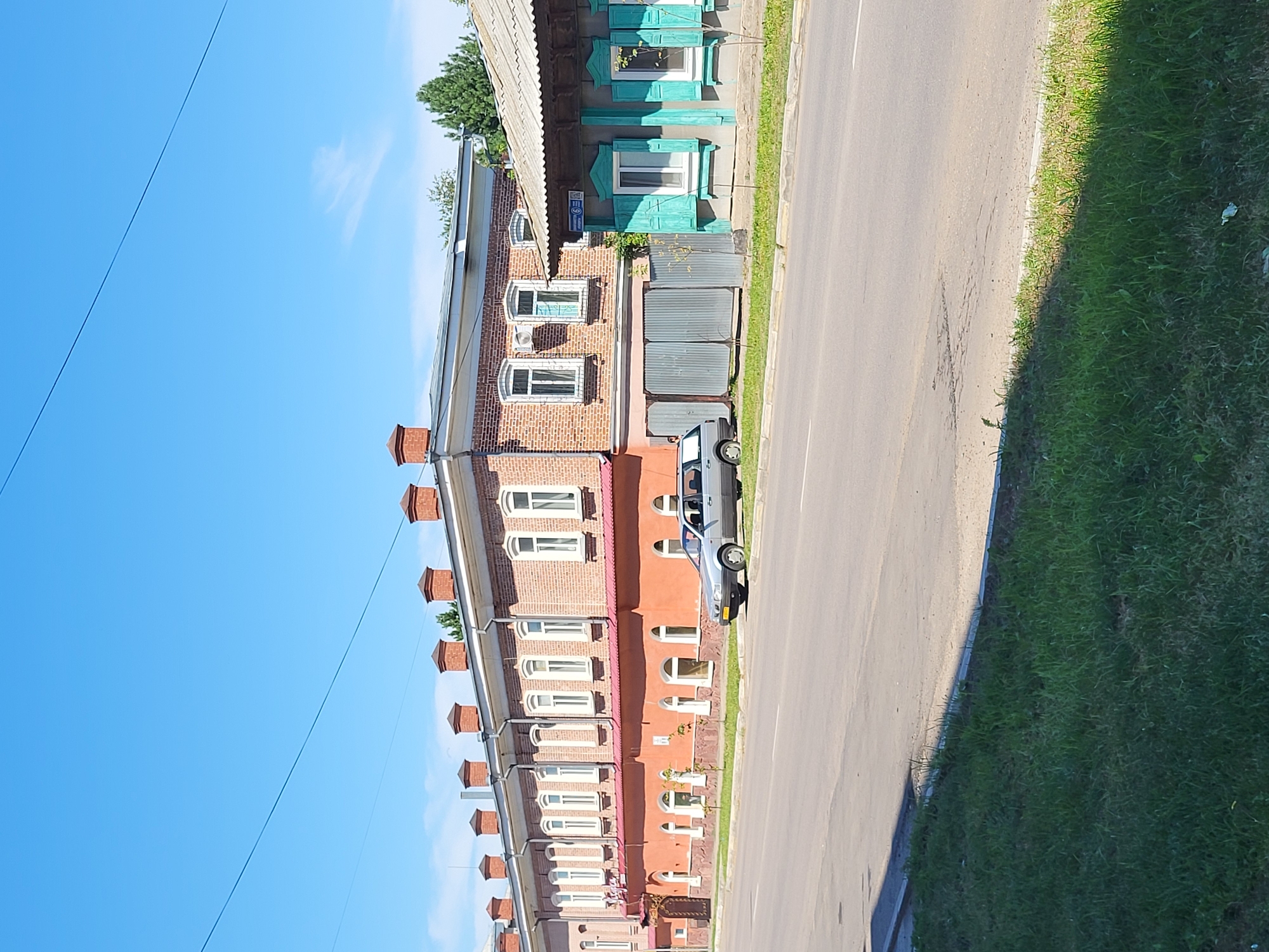 A street view shows a light gray station wagon parked on the right side of an asphalt road. The car is positioned in front of a long, two-story brick building with an orange lower facade and white-trimmed windows. This building has a red roofline with numerous brick chimneys. To the right of the brick building, a smaller wooden structure with turquoise shutters and a corrugated metal roof is visible. A green grassy area borders the road in the foreground, with a faint shadow cast across it. The sky above is clear and blue. The location is identified as the city of Petropavl, Kazakhstan, on a street named Mira Street, as indicated by a street sign.