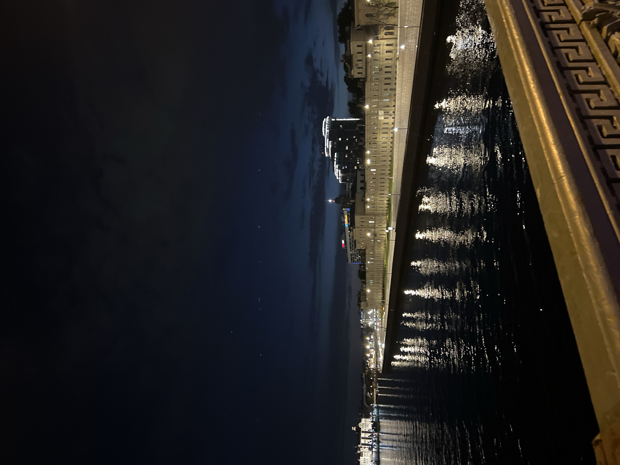 A night scene in Saint Petersburg, Russia, viewed from a bridge. The sky is dark, with hints of clouds. Along the water's edge are buildings illuminated by streetlights. The lights reflect on the dark water. A metal railing, adorned with a Greek key design, is visible on the right side of the frame.