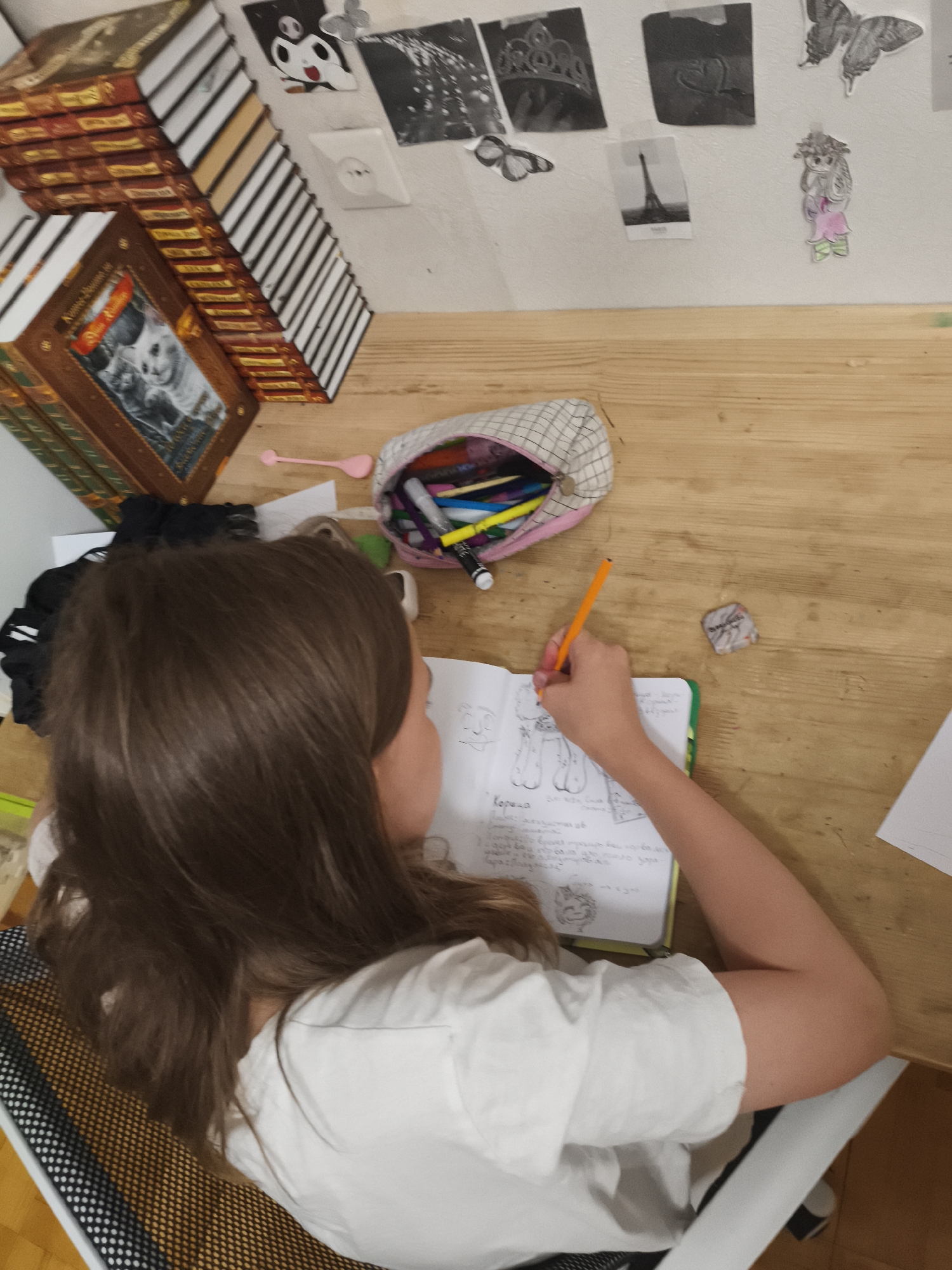 An overhead photograph captures an individual sitting at a wooden desk in an indoor setting, observed from behind their right shoulder. The person, with long, medium-brown hair, wears a white short-sleeved t-shirt and is seated in a chair with a black and light-brown checkered mesh backrest. Their right hand holds an orange pencil, actively drawing or writing in an open, spiral-bound notebook with a green spine. The notebook displays sketches of figures and handwritten text in Cyrillic, with the word "Корона" (Korona) visible on the lower left page.
The wooden desk surface contains several items. To the upper left of the notebook is a light-colored, patterned pencil case filled with various colored markers and pencils, one black marker with a yellow cap partially extended. A small, pink, spoon-shaped object lies to the left of the pencil case. A crumpled piece of paper is situated to the upper right of the notebook, and a blank white sheet of paper rests on the far right edge of the desk.
On the left side of the desk, a stack of approximately 10-12 books with brown, gold, and reddish spines is arranged, some horizontally, some vertically. The most prominent book cover features two cats (one white, one dark) and the Cyrillic title "Коты-Воители" (Warrior Cats). Behind this stack, another stack of approximately 15-20 books with white spines and black horizontal stripes is diagonally arranged against the back of the desk.
The white wall behind the desk is decorated with several taped-on images and cutouts. These include: a black and white cutout of a cartoon character's head (white with black features and two red cheek spots, resembling a frog-like character); a small gray butterfly cutout; a black and white print of a crown/tiara; a black and white print featuring a heart shape; a black and white print of the Eiffel Tower labeled "Paris"; another butterfly print; and a cutout of a cartoon-style girl with light hair, a pink skirt, and a green top. A white electrical outlet with a single circular European-style socket is visible on the wall below the cartoon character and butterfly. The scene is evenly lit, indicating an indoor environment in the city of Unknown, Unknown.