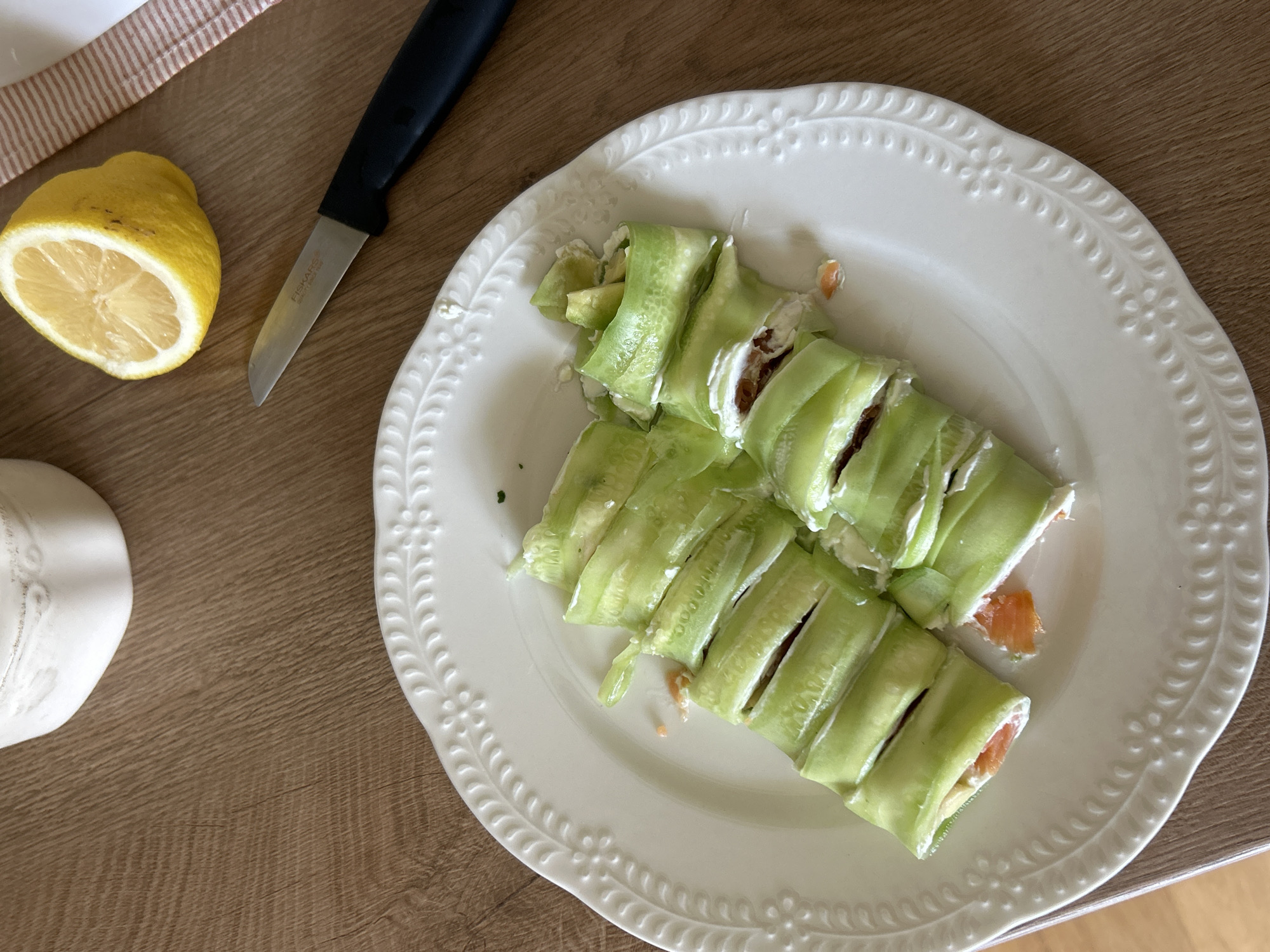 A white, embossed ceramic plate containing cucumber rolls is centered on a wooden table. The rolls are made from thinly sliced cucumber wrapped around a filling, possibly cream cheese, with visible pieces of salmon and avocado. A partially eaten lemon and a black-handled knife are positioned on the table to the upper right of the plate. A white ceramic pitcher is visible in the upper left corner. The lighting suggests an indoor setting, possibly a kitchen or dining area. There is no indication of the city of Plovdiv, Bulgaria.