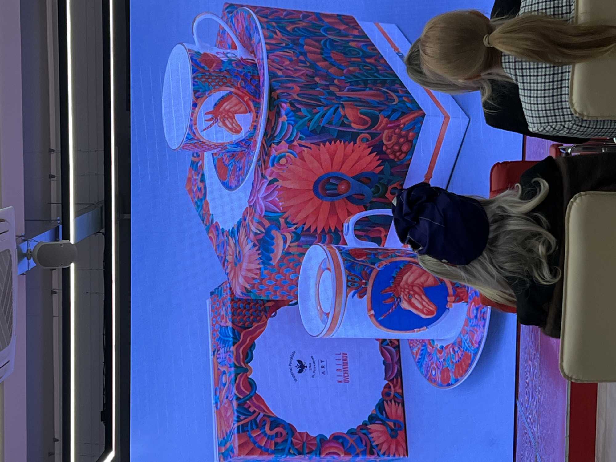 The image shows a display featuring porcelain products, likely tea cups and boxes, designed by Kirill Ovchinnikov Art from Imperial Porcelain, Saint Petersburg, Russia. The products are decorated with vibrant, detailed floral and mythical creature designs. Two individuals are partially visible: one with blonde hair pulled back in a ponytail wearing a checkered jacket, and another with long, light blonde hair wearing a dark-colored outfit and a navy blue hat. They appear to be observing the display. The display itself is lit with edge lighting. The scene seems to be inside a showroom or exhibition space.