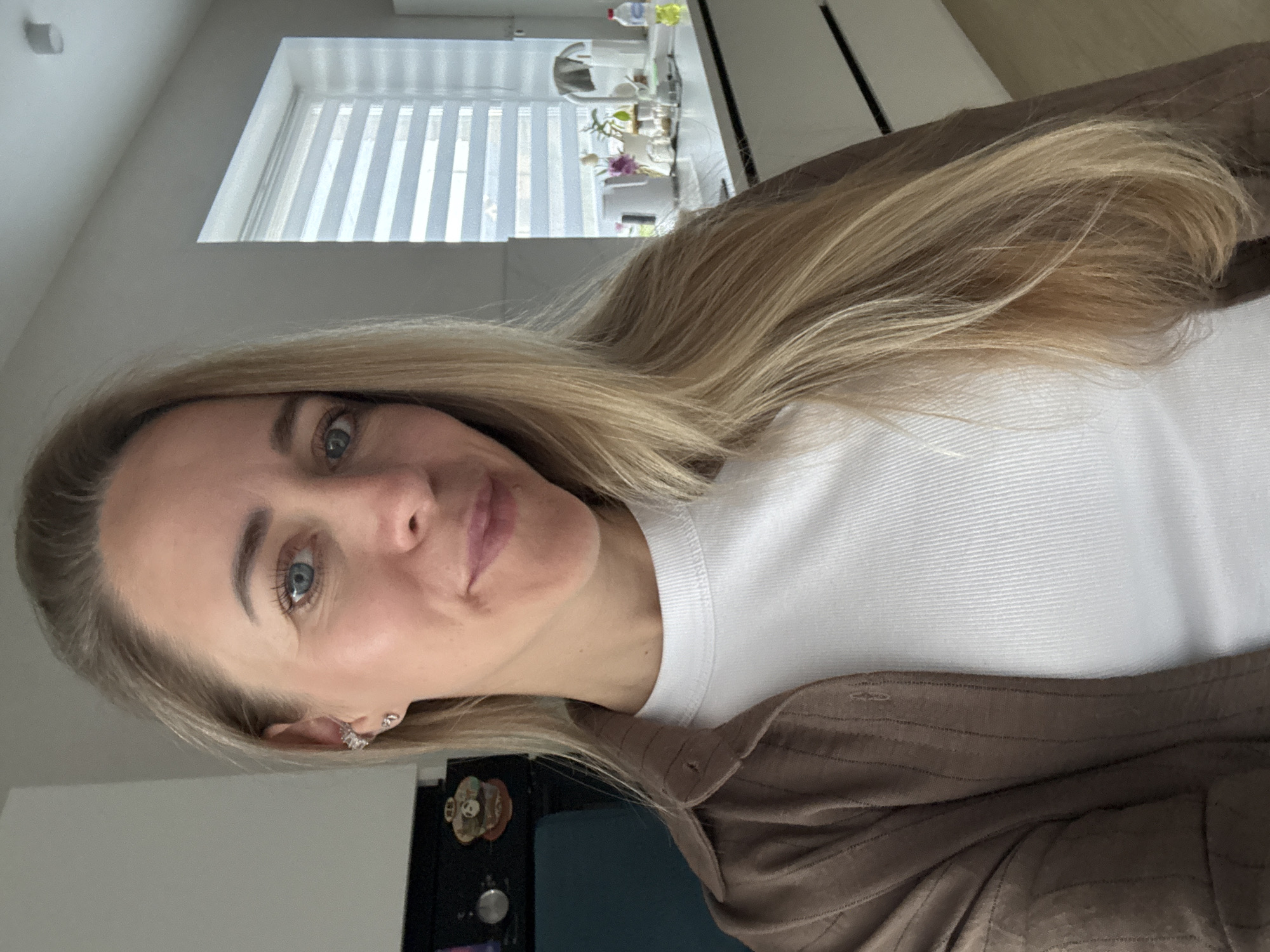 A woman with blonde hair and blue eyes is in a kitchen. She is wearing a white t-shirt and a brown shirt. There is a window with blinds behind her, and a countertop with kitchen appliances. There is a black object on the wall to her left. The woman is looking at the camera. The image appears to be a selfie. The location is Chaikovsky, Russia.