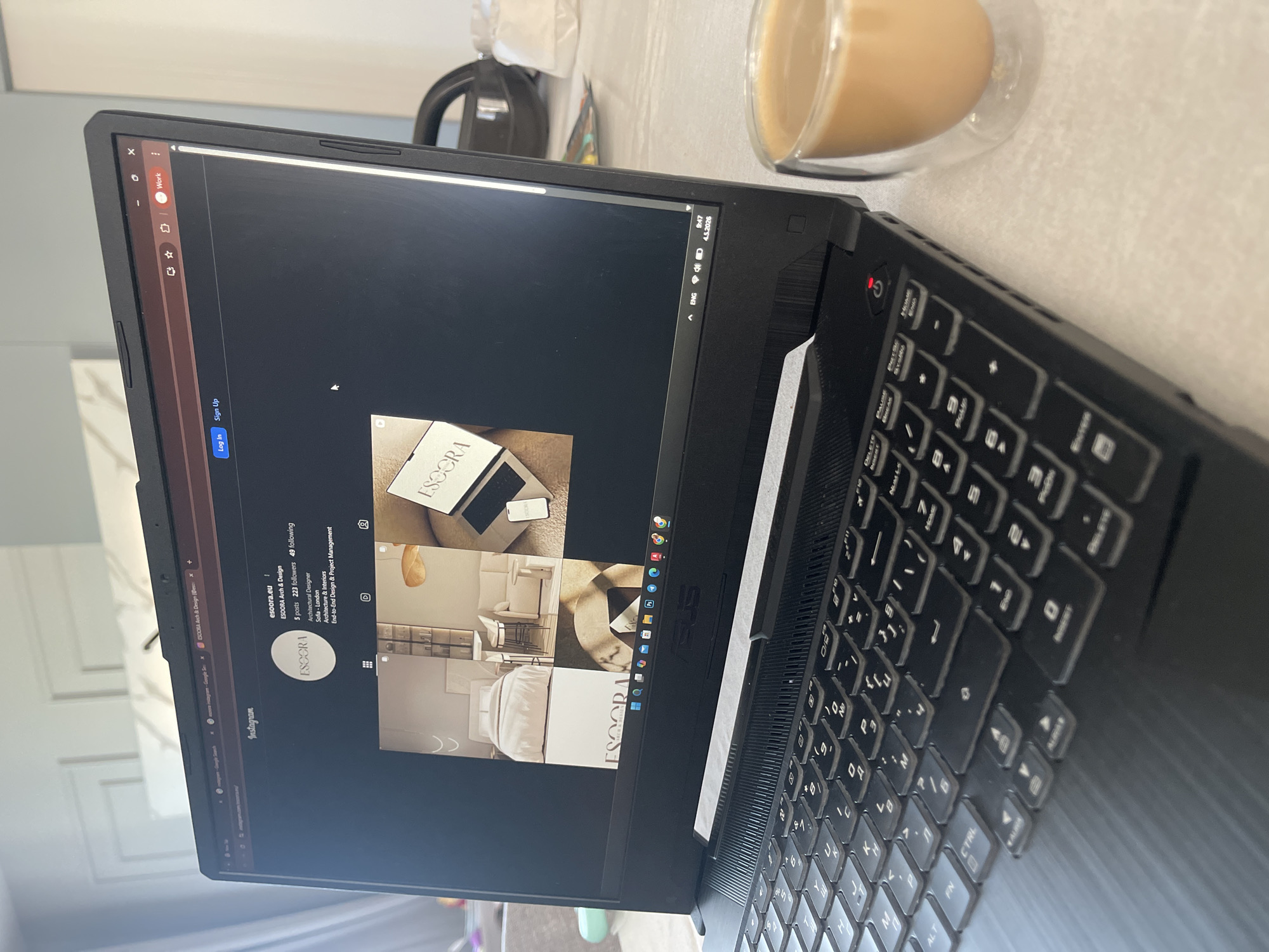 The image shows a black ASUS laptop open, displaying the Instagram profile of "escora.eu - ESCORA Arch & Design". The laptop is positioned on a light-colored surface, possibly a table, next to a clear glass mug filled with a beige liquid, likely coffee or tea with milk. A black object is visible blurred in the background, possibly a kettle. The laptop keyboard is partially visible, angled towards the right side of the frame. The timestamp on the laptop indicates "9:47 4.5.2026". The scene is indoors.