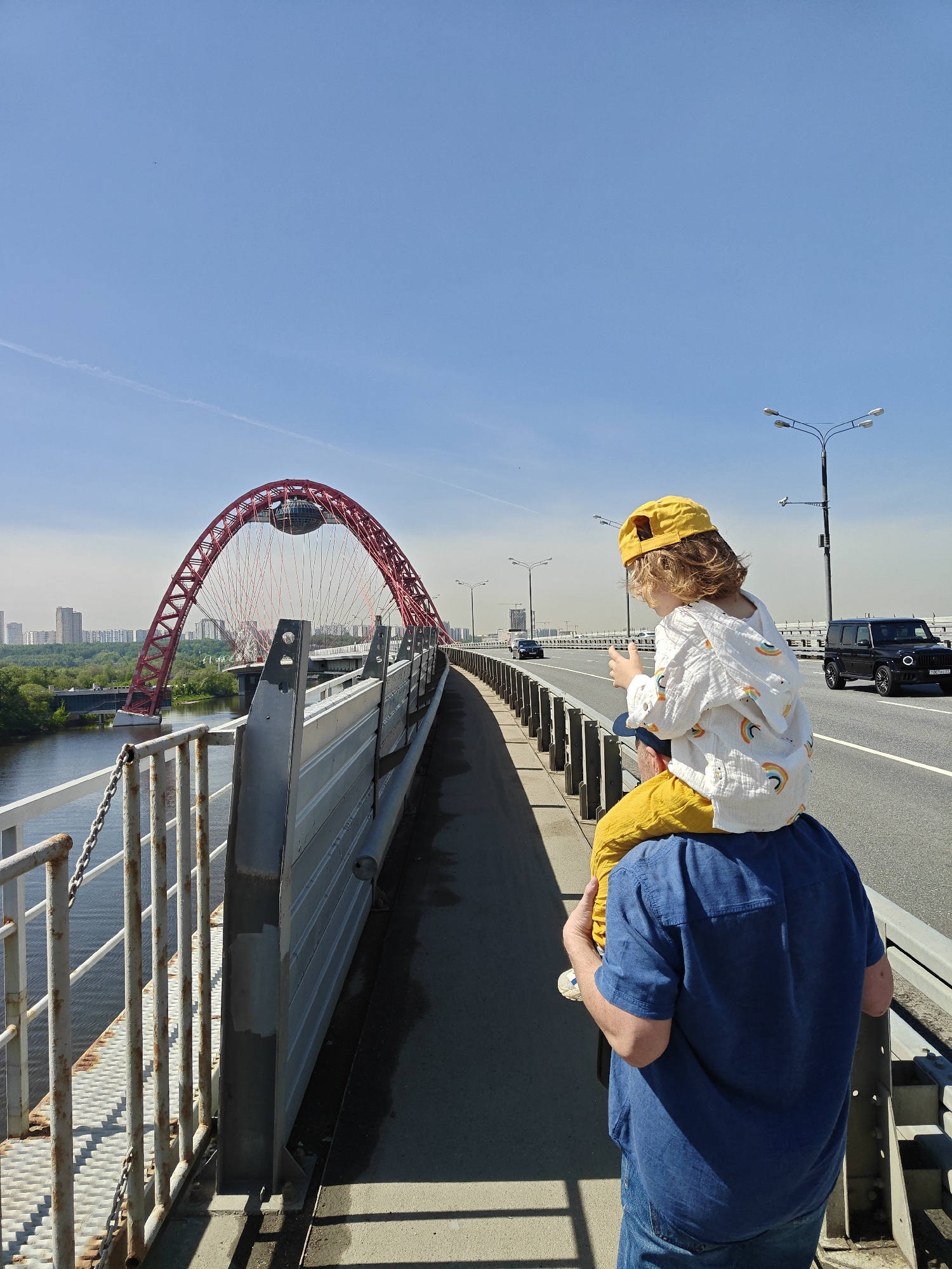 A medium eye-level outdoor shot shows the back of a man with a child riding on his shoulders, walking along a gray pedestrian walkway on a bridge in Moscow, Russia. The man wears a short-sleeved blue shirt and denim jeans; the child wears a yellow baseball cap, a white jacket with rainbow images, and mustard-colored pants. The walkway, positioned on the left side of the bridge, is separated from the multi-lane traffic by a corrugated guardrail. To the left of the walkway, a chain-link fence runs along the edge of the bridge, overlooking a body of water. The Zhivopisny Bridge, a cable-stayed bridge with a distinctive red arch, spans the river in the background. On the right side of the traffic, street lamps stand along the highway. A black SUV is seen traveling on the highway. In the far background, a skyline of buildings is visible. The sky is clear and blue.