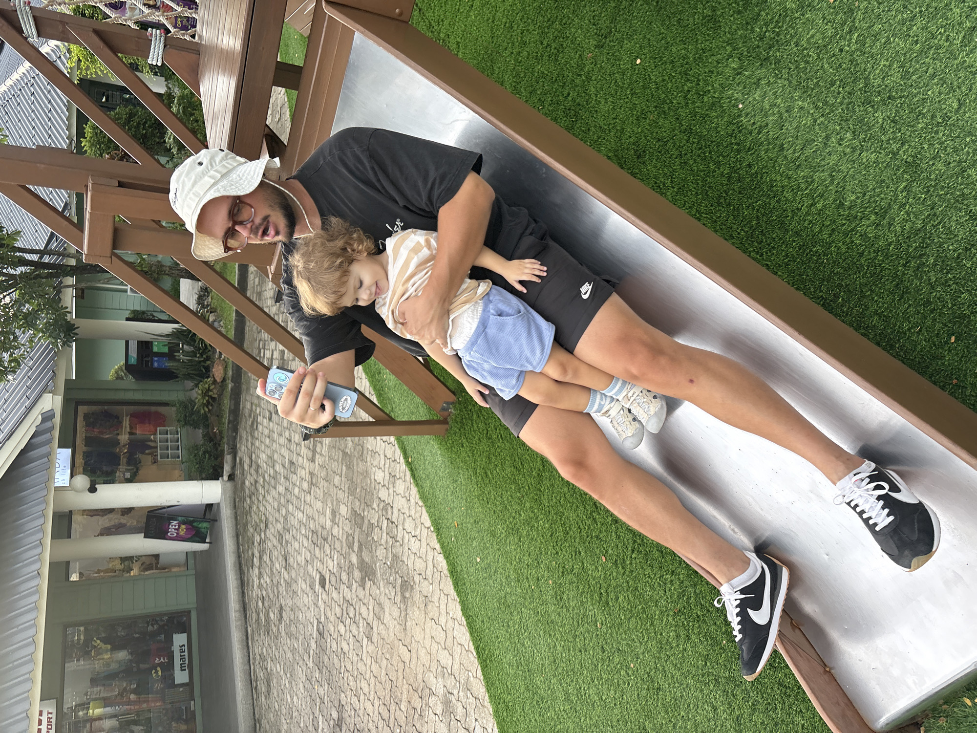 A man wearing a white bucket hat, glasses, and a black t-shirt sits on a metal slide, holding a young child. The child wears a striped shirt and blue shorts. The man is holding a smartphone in his right hand, appearing to take a selfie. The slide is part of a wooden playground structure, set against a backdrop of artificial turf and paved ground. In the background, a building with shopfronts is visible, with signage that appears to be in English. The overall setting suggests an outdoor recreational area, possibly a park or a resort, located in the city of Choeng Thale, Thailand.