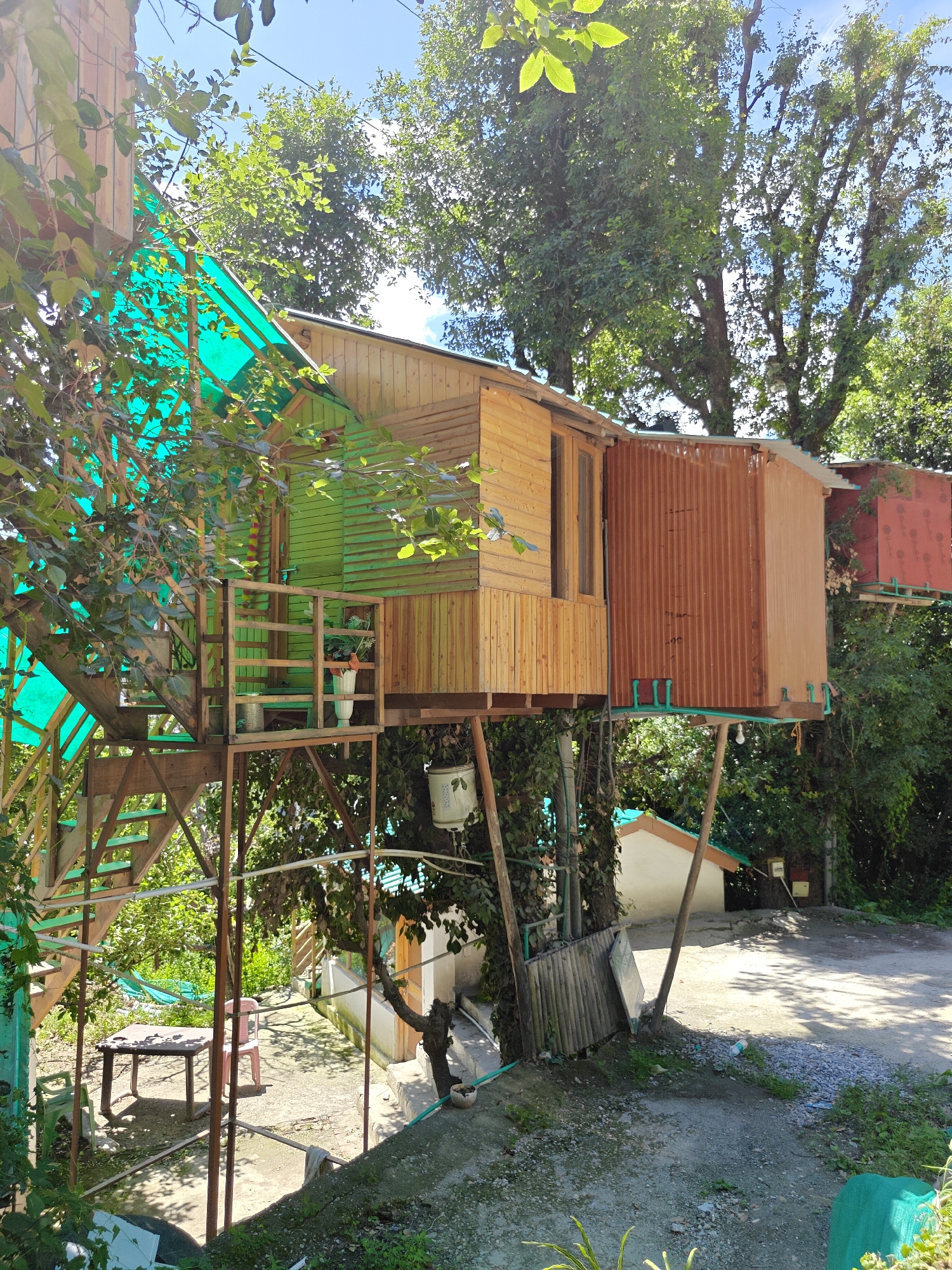 This is a photograph of a treehouse complex located in Prini, India. The structure is elevated and made of wood, with a pitched roof and wooden siding. A bright green tarpaulin covers part of the structure and stairs leading up to it. There are multiple levels and sections to the treehouse, with visible wooden stairs and railings. The treehouse is surrounded by lush green trees and foliage, suggesting a natural, outdoor setting. The ground level features a concrete or paved area with a wooden table and chairs, indicating an outdoor seating or dining area. The overall scene is one of rustic charm, likely a guesthouse or tourist accommodation.
