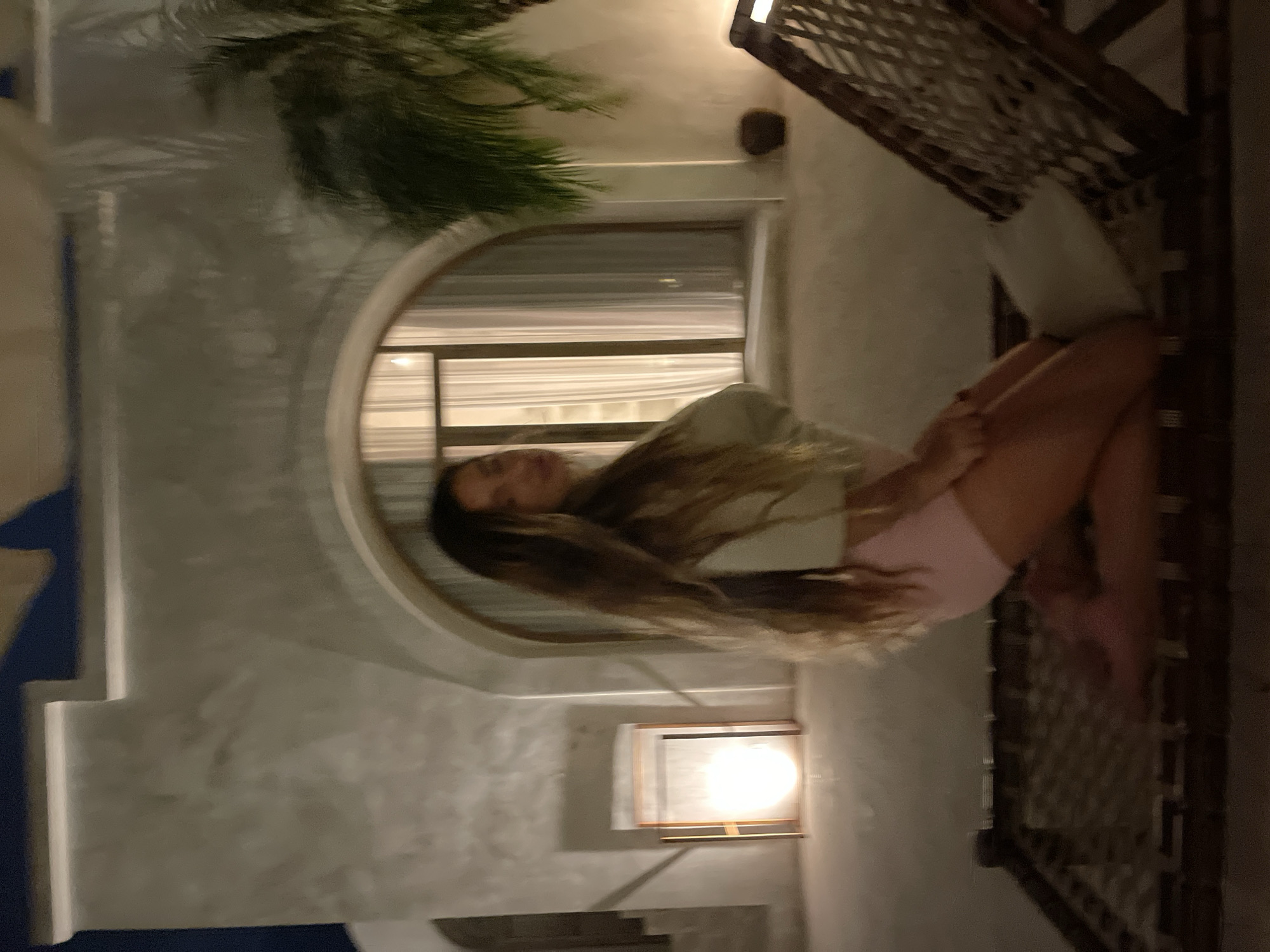 In an outdoor evening setting, a young woman with long, light brown hair kneels on a woven cot. She is wearing a light-colored top and pink shorts. Her body is positioned facing the left side of the frame, with her head turned slightly towards the viewer. Behind her, a large arched window is illuminated from within by sheer curtains. To the left of the window, a tall, rectangular lamp with a wooden frame casts a bright light. The background features a textured, light-colored wall, and to the right, a palm tree is visible. The ground appears to be a light-colored, possibly sandy or paved surface. A wicker chair with a white cushion is positioned in the foreground to the right of the woman. The lighting suggests a warm, ambient atmosphere, possibly from artificial sources and the setting sun.
