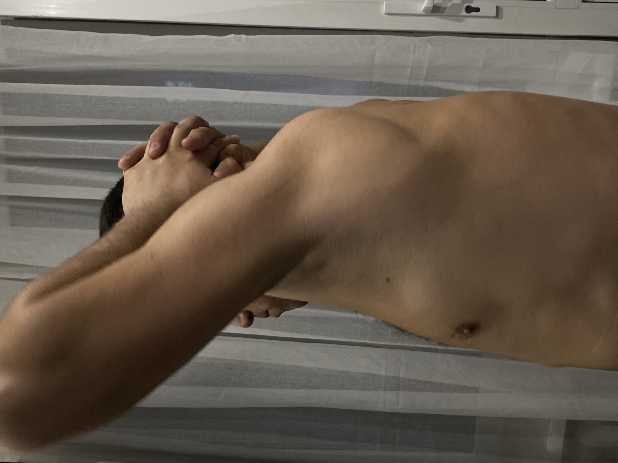 A person is seen shirtless in a photograph. The person is light-skinned with dark hair. Their hands are clasped together near their head, which is bowed. The background consists of a window with light-colored blinds. The image provides no information about the city of Sochi or any other location.