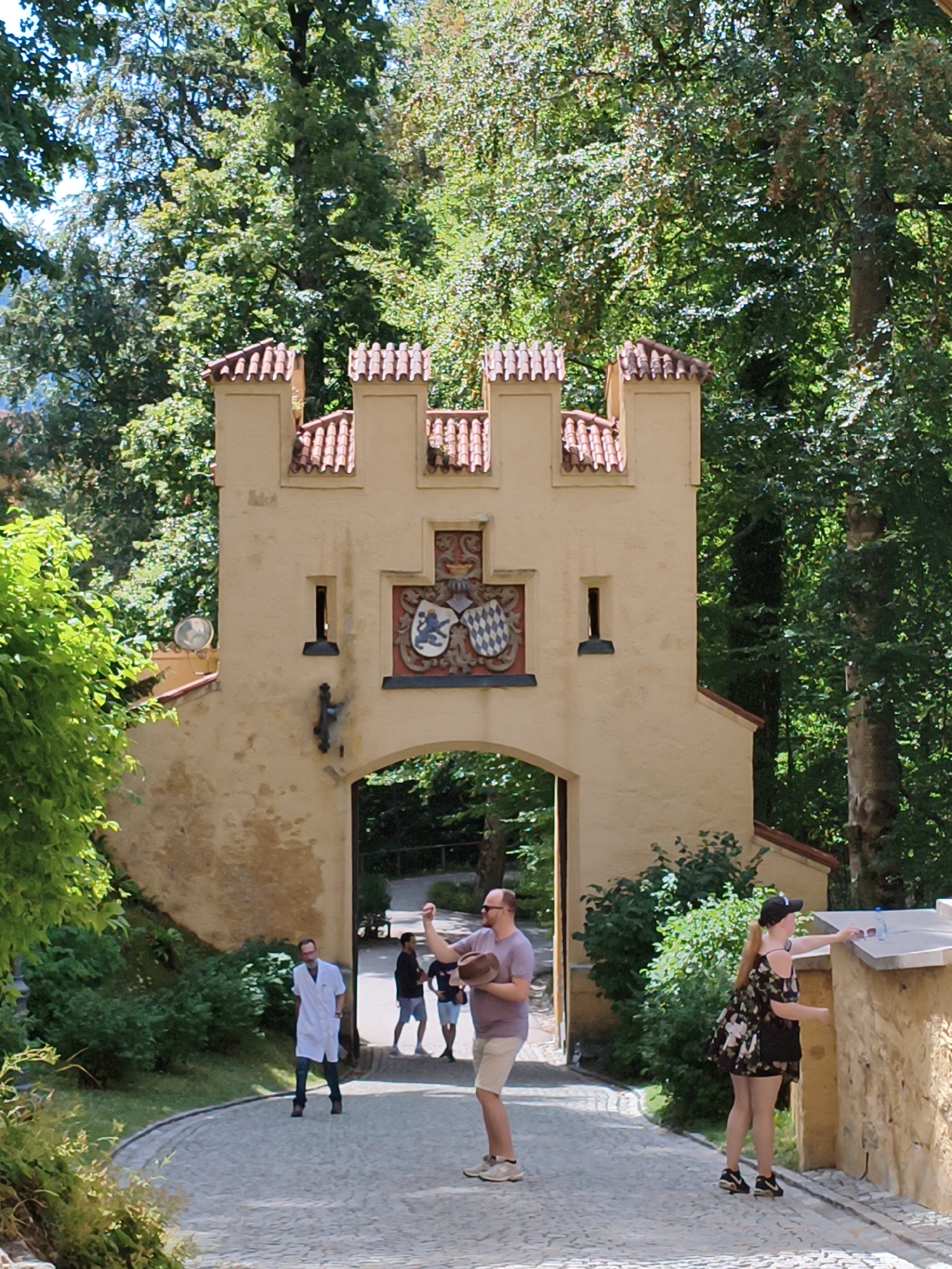 The image shows a castle gate in Schwangau, Germany, featuring a cream-colored facade with a red-tiled roof. The gate has an arched entryway, and above it, a crest with two shields is displayed. The area surrounding the gate is lush with green trees and foliage. Several people are present: a man in a white lab coat walks towards the gate; another man in a purple shirt and shorts holds a hat; and a woman in a floral dress and a black cap is reaching for a pair of sunglasses on a stone wall. The path leading to the gate is paved with cobblestones.