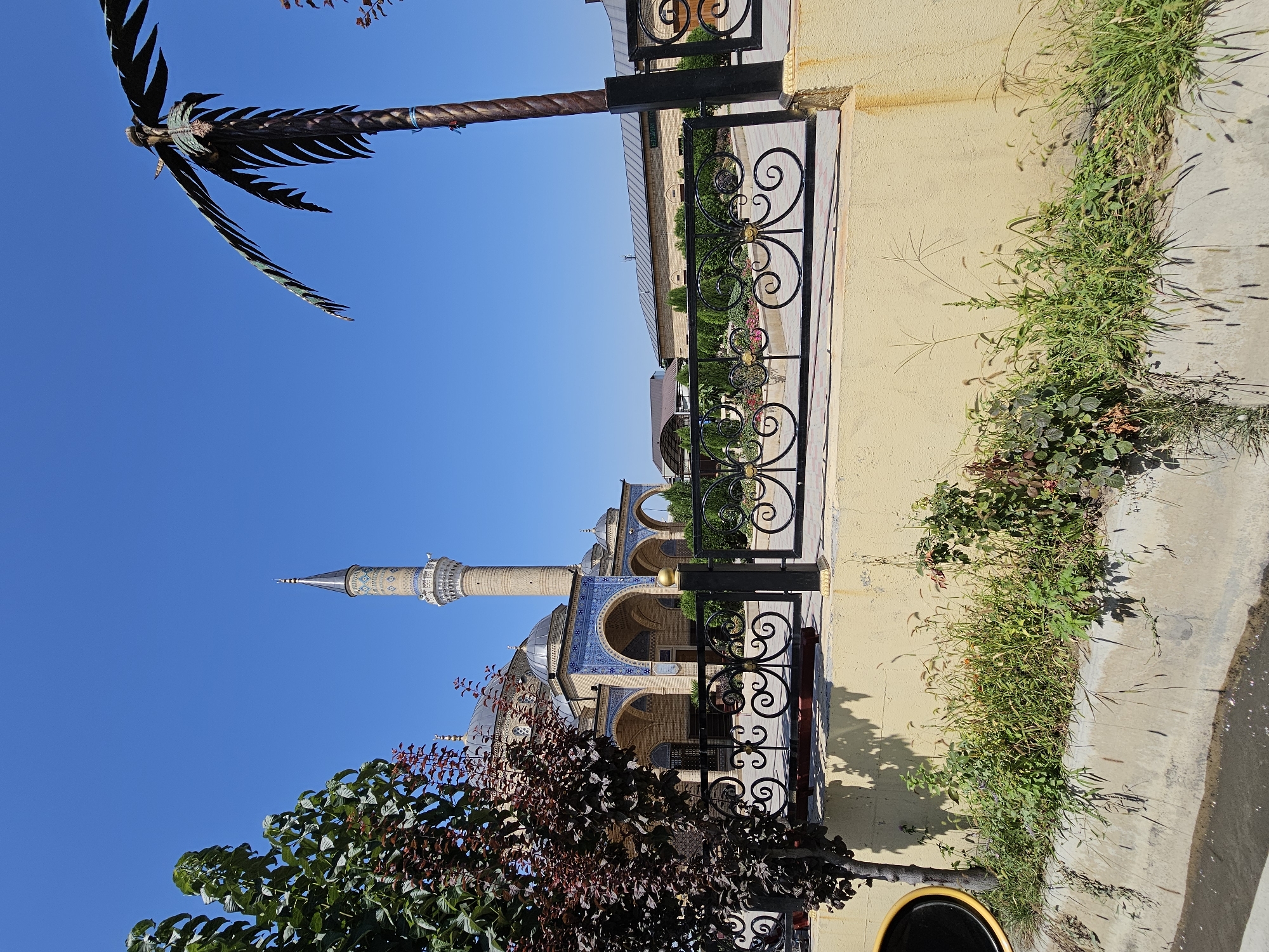 The image depicts the exterior of a mosque in the city of Özgön, Kyrgyzstan, under a clear blue sky. In the foreground, a concrete channel runs along the bottom of the frame, with overgrown green vegetation and some scattered brown grasses growing on the bank above it. A light yellow wall forms the upper bank of the channel. A black, decorative metal fence with ornate scrollwork and gold accents runs horizontally across the mid-ground. To the left of the fence, a tree with dark reddish-brown leaves is partially visible. Behind the fence, the mosque complex is visible. The mosque itself has a light brown brick facade and domes. A tall, slender minaret with intricate geometric patterns in shades of blue and white rises from the mosque complex, reaching towards the sky. Arched walkways with blue tiled accents surround a portion of the mosque. To the right of the minaret, a decorative metallic palm tree sculpture stands tall against the blue sky. In the background, other buildings with light brown brick construction and sloping roofs are visible, along with manicured green bushes and trees. The overall scene is one of a peaceful, well-maintained religious site in an urban environment.