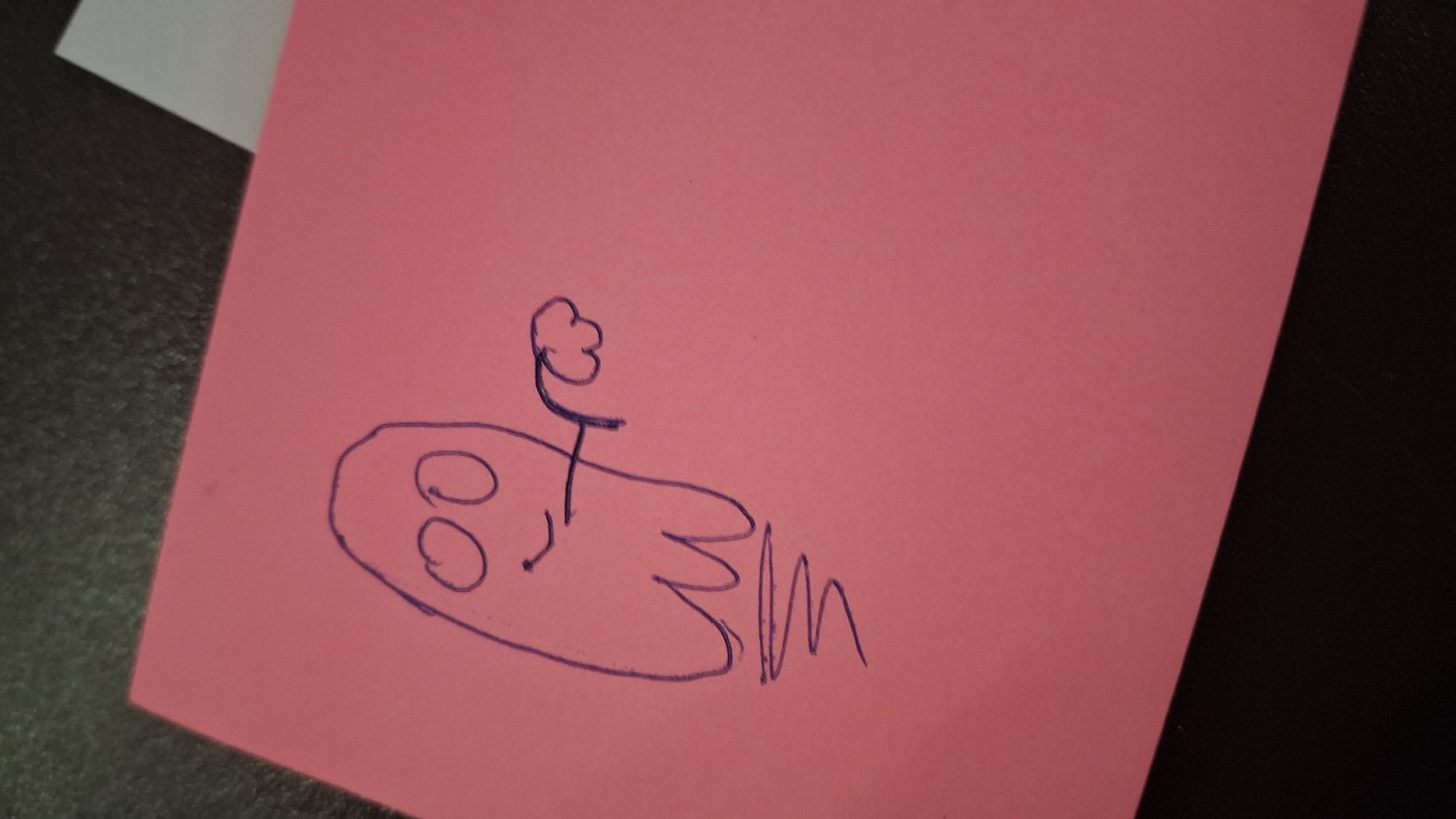 A hand-drawn purple ghost with wide eyes, a smiling mouth, and a wisp of smoke or mist coming from its mouth is depicted on a pink rectangular surface. The ghost is floating and has its right arm extended with a puffy, cloud-like shape at the end of it. Below the ghost, there are several wavy lines, possibly representing its spectral trail or base. The pink surface appears to be a piece of paper or a sticky note, and it is placed on a dark, textured surface. The background is out of focus and shows a dark area and a lighter area, possibly indicating a table and a wall or another object. The image is a close-up shot, with the ghost drawing occupying the left and central portion of the frame.