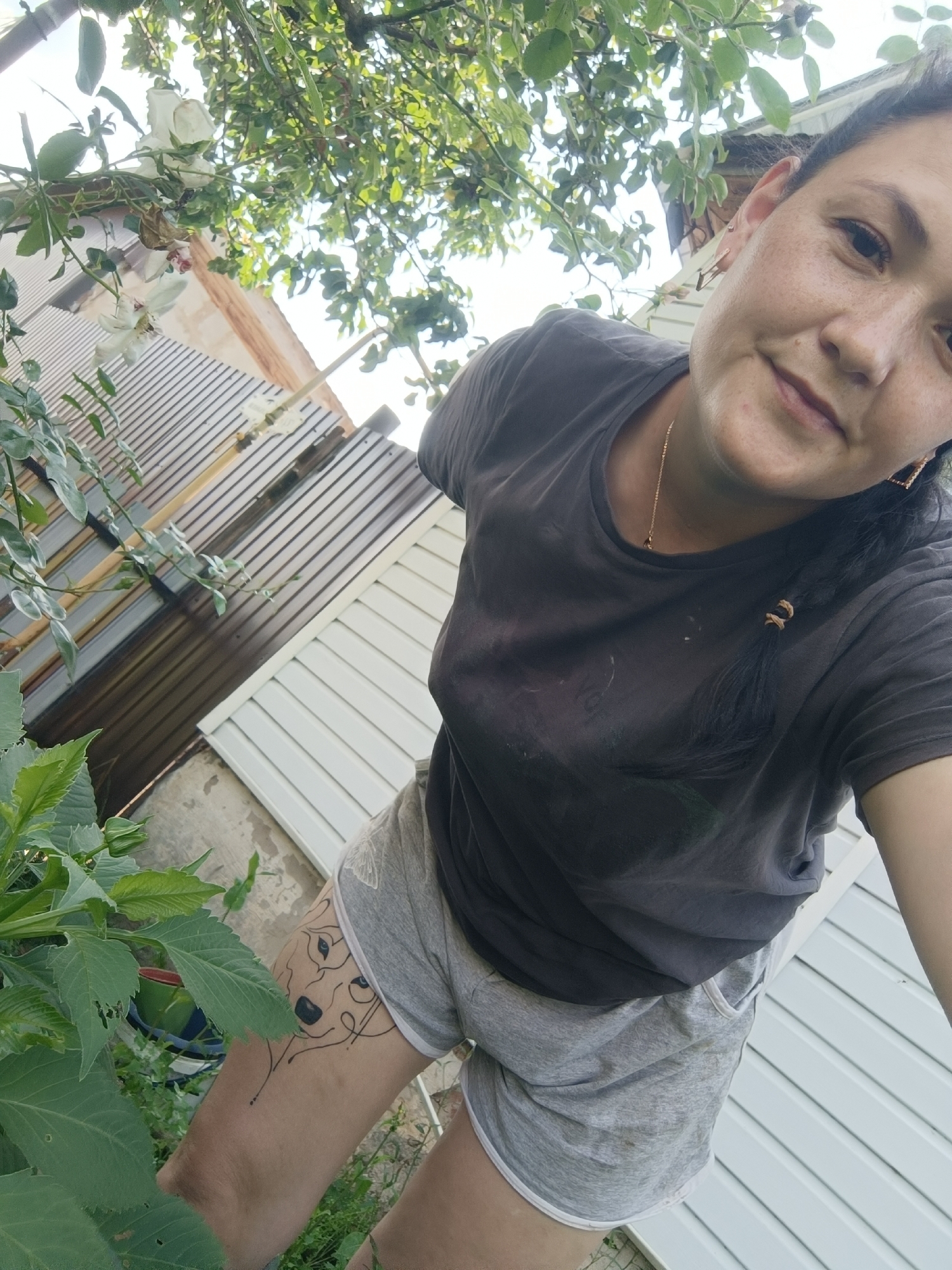 In Karsha, Russia, a woman with dark hair braided and wearing a dark grey t-shirt and light grey shorts poses outdoors. Her left thigh features a black line drawing tattoo of a wolf's face. She is positioned near a lush green plant. In the background, a fence with a brown corrugated metal section and white siding is visible, alongside a tree with white roses and green leaves overhead. The overall scene suggests a residential outdoor setting.