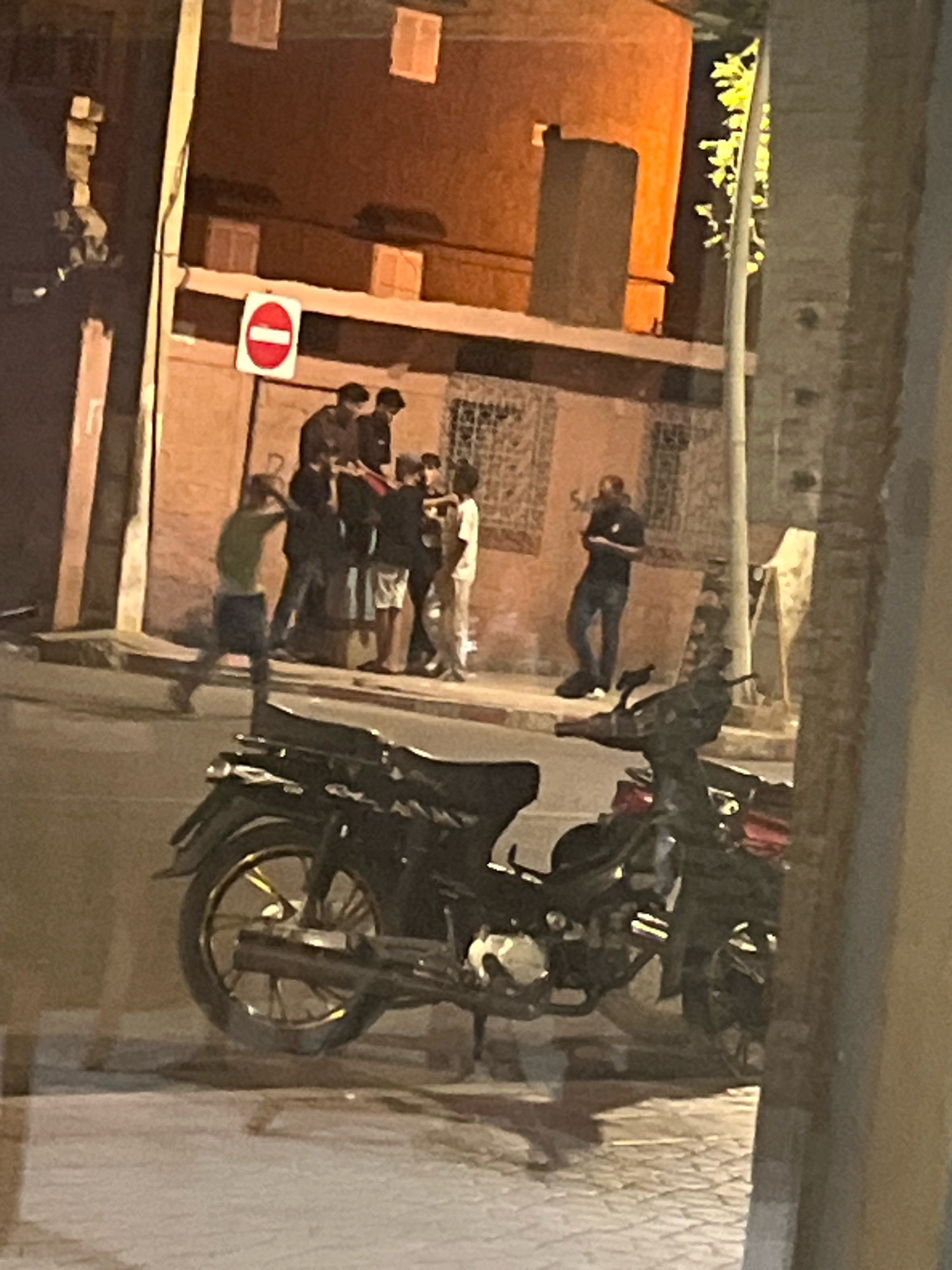 The image depicts a street scene at night in Azilal, Morocco. In the foreground, two motorcycles are parked, one in front of the other, partially obscuring the view of the street and sidewalk. The motorcycles are dark in color, with visible wheels and engines.
In the background, a group of individuals, appearing to be young men, are gathered on the sidewalk in front of a building. One individual in a green shirt is in motion, running away from the group, with his arms outstretched. The other individuals are standing close together, some looking towards the person running, while others are facing away. They are dressed in casual clothing.
The building behind them has a reddish-orange facade with visible windows. A red and white "do not enter" sign is posted on the wall above the group. The street is paved with cobblestones. The lighting suggests artificial streetlights and illumination from within the building. The overall atmosphere appears to be a moment captured in time, possibly during an interaction or event.