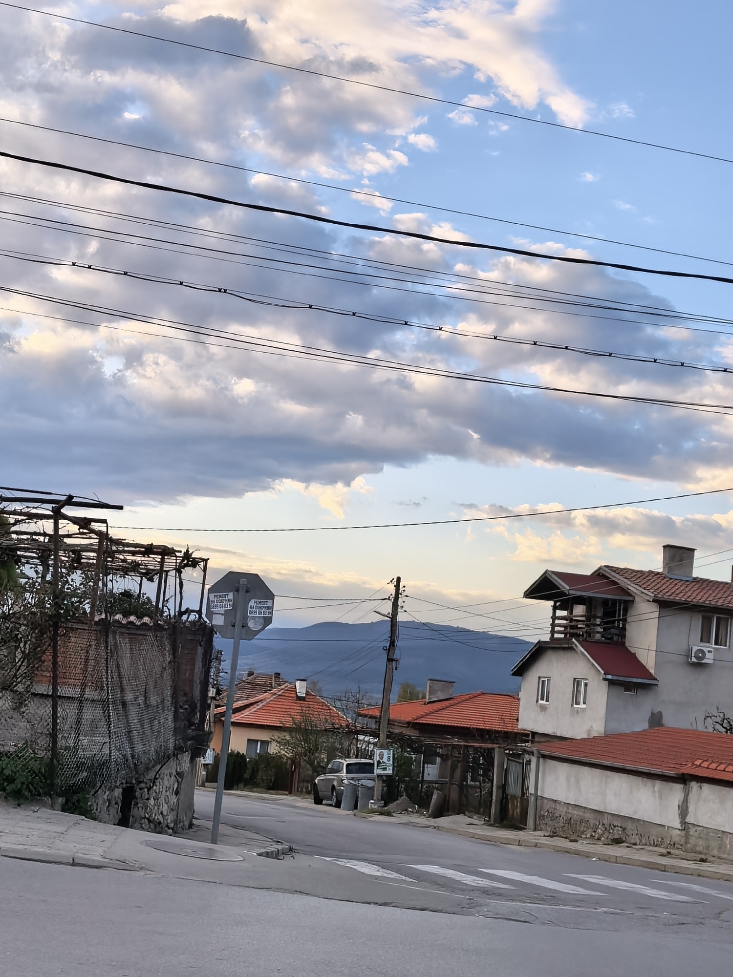 The image shows a street scene in an urban area, likely a town or village. The sky is partly cloudy with blue patches visible. Overhead are several electrical or utility wires.
On the left side of the street, there is a partially enclosed structure made of brick, stone, and wire mesh. A sign attached to a pole is visible, with writing in a non-English language. The street slopes gently downhill with a crosswalk painted on the road surface.
On the right side of the street are several buildings with red tiled roofs. One building has a second-story addition with a balcony. An air conditioning unit is attached to the side of this building. A telephone pole is located near the buildings.
In the distance, a mountain range is visible under the clouds. A car is parked further down the street near some signage. The scene appears to be a residential area on a quiet street.