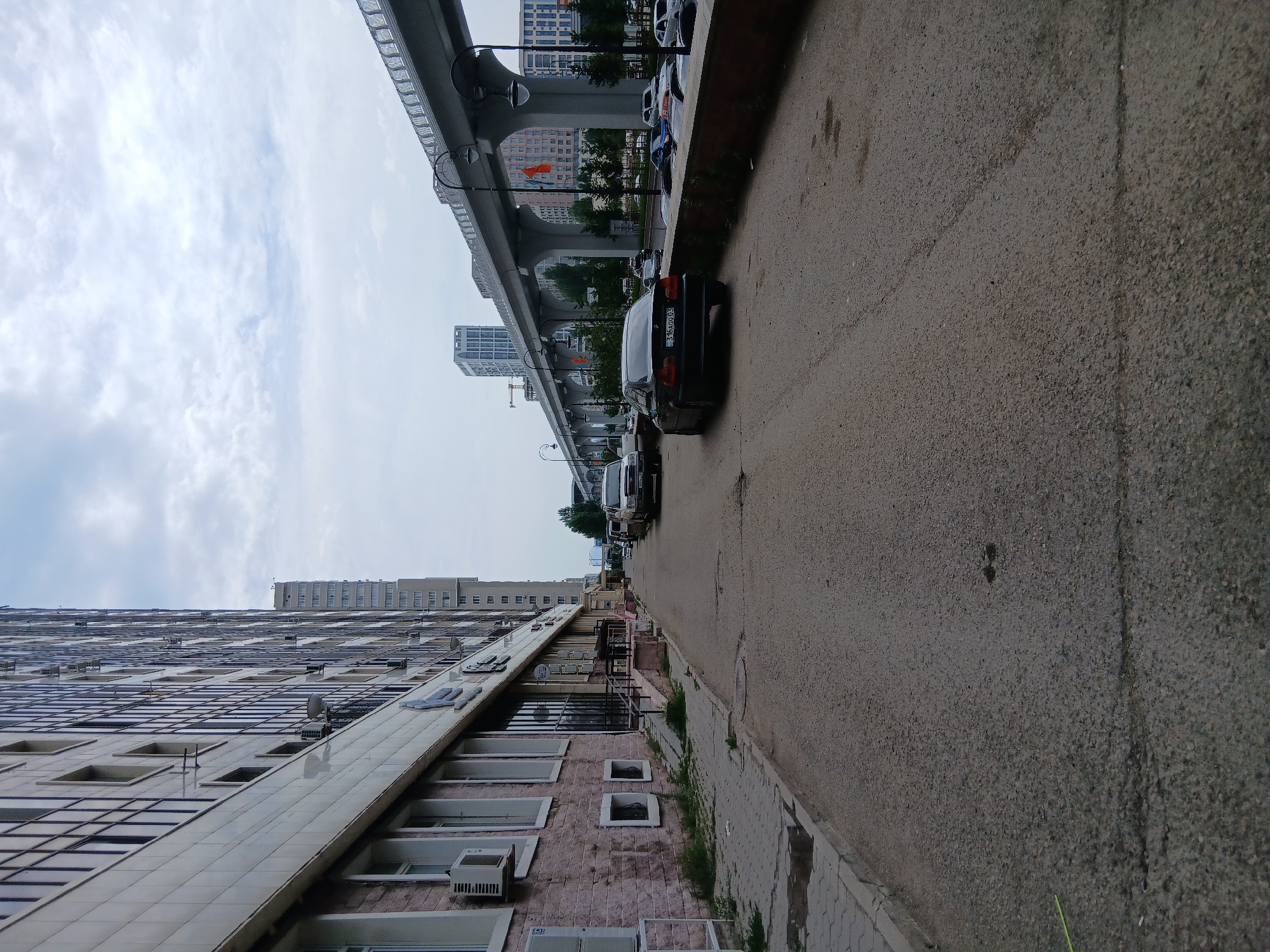 The image depicts a street scene in Astana, Kazakhstan, with a prominent modern high-rise building on the left. The building's facade is characterized by numerous windows and external air conditioning units. A paved sidewalk runs alongside the building, showing signs of wear, including puddles and some overgrown grass. The street itself is asphalted and appears to be a public road.
Several vehicles are visible on the street. A dark sedan is parked on the right side of the road, facing away from the camera. Further down the street, other vehicles are parked or in motion, including a white van. A raised concrete overpass, likely part of a transportation infrastructure like a monorail or elevated highway, dominates the background, stretching across the scene and supported by large concrete pillars. Beyond the overpass, more modern buildings and structures are visible, some with orange flags displayed. The sky is overcast with a mix of clouds. The overall impression is of a contemporary urban environment.