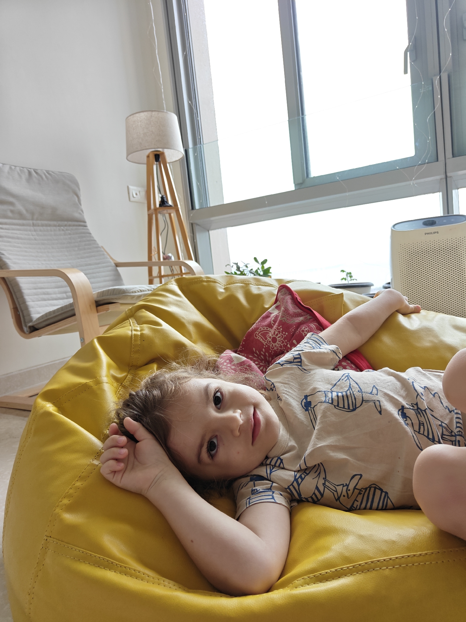 A young child with wavy brown hair lies on a bright yellow beanbag chair, looking upwards and to the left. The child is wearing a beige t-shirt with a blue line drawing of a whimsical creature. A red and white patterned pillow is partially visible behind the child. The beanbag is in a sunlit room with a large window that offers a view of the sky. To the left, a grey and wooden rocking chair is positioned. In the background, a floor lamp with a cream-colored shade and wooden legs stands near the window. On the right side of the window, a white Philips air purifier is visible. The overall environment suggests a modern, comfortable living space. The image does not provide specific clues to identify the location as Mumbai, India.