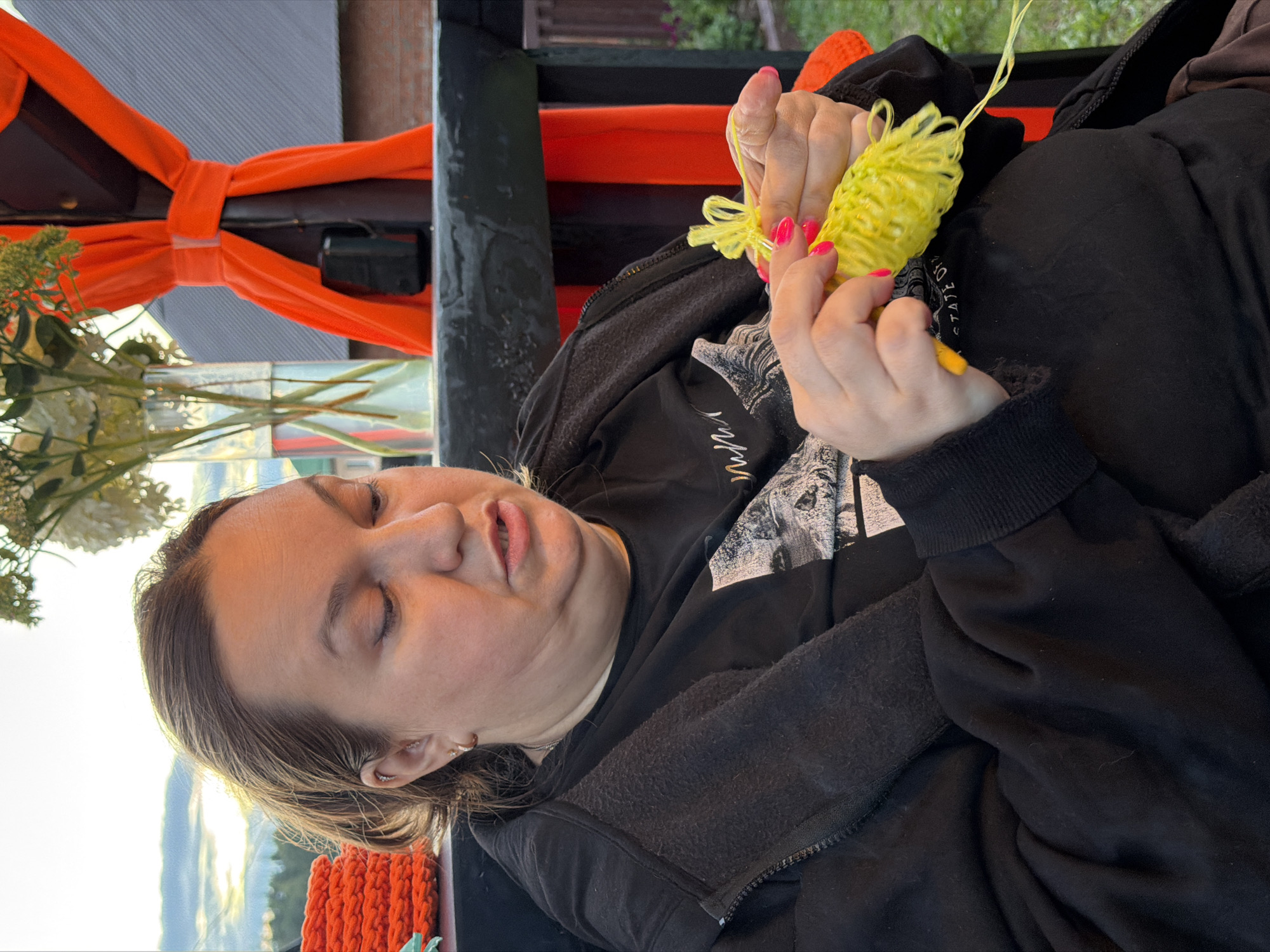 A medium shot captures a woman with blonde hair pulled back, wearing a black zip-up jacket and a black t-shirt, as she engages in knitting with bright yellow yarn. Her hands, adorned with bright pink nail polish, are actively manipulating the yarn. The background suggests an outdoor setting with a black railing, an orange draped fabric, a vase of flowers, and a glimpse of a wooden fence and buildings. The lighting indicates it's either early morning or late evening, given the soft, warm glow and the presence of clouds in the sky. The woman's facial expression is one of concentration. The location is identified as Chaikovsky, Russia.