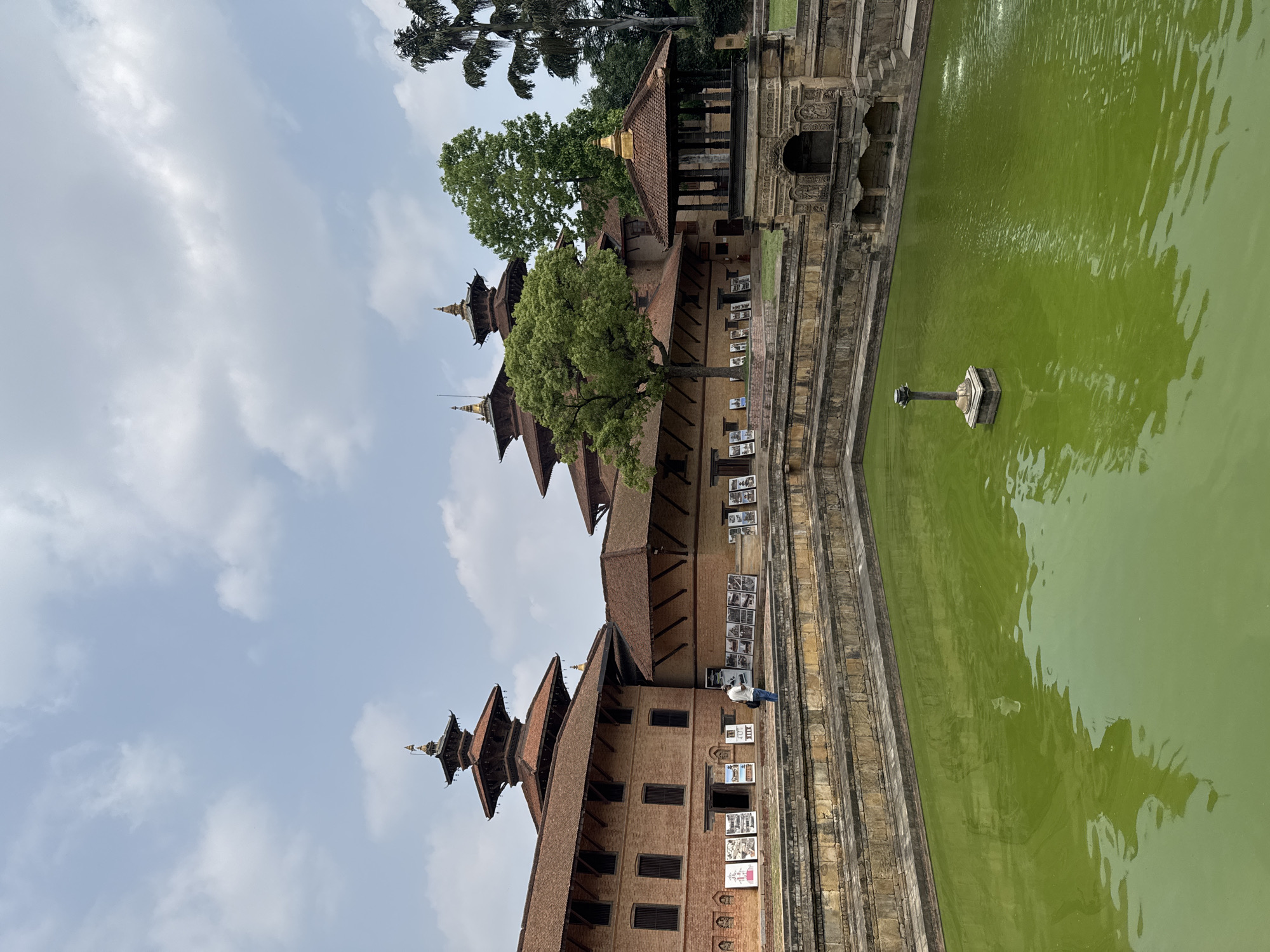 The image shows Patan Durbar Square in Lalitpur, Nepal. A multi-tiered pagoda-style building is on the left, constructed of brick and wood. The building displays numerous photos and signs affixed to its facade. A man stands beside the building near a doorway. A large rectangular pond is next to the building with greenish water and a central fountain or column. A stone staircase leads down to the pond's edge. The sky is visible above the building, showing light clouds. A tree is in the upper center, and another partially visible tree is at the top.