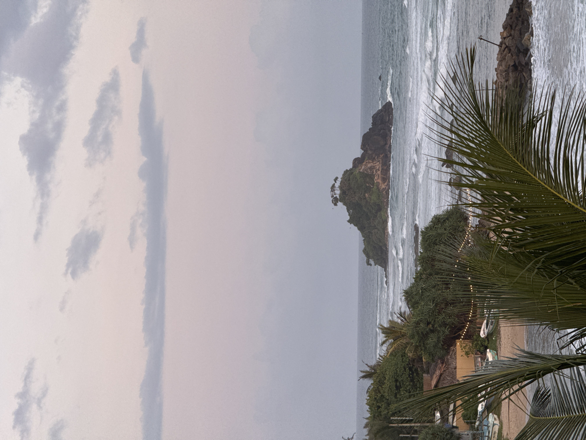 A scenic view captures a tranquil beach scene at Goviyapana, Sri Lanka. The foreground is dominated by the fronds of a palm tree, framing a sandy beach dotted with several small boats. String lights are visible, strung along a fence that separates the beach from lush green vegetation. Beyond the beach, the turquoise ocean stretches out, with gentle waves lapping at the shore. In the distance, a small, rocky island rises from the water, crowned with a cluster of trees, its silhouette softened by the hazy, pastel-colored sky. The overall atmosphere is serene and picturesque.
