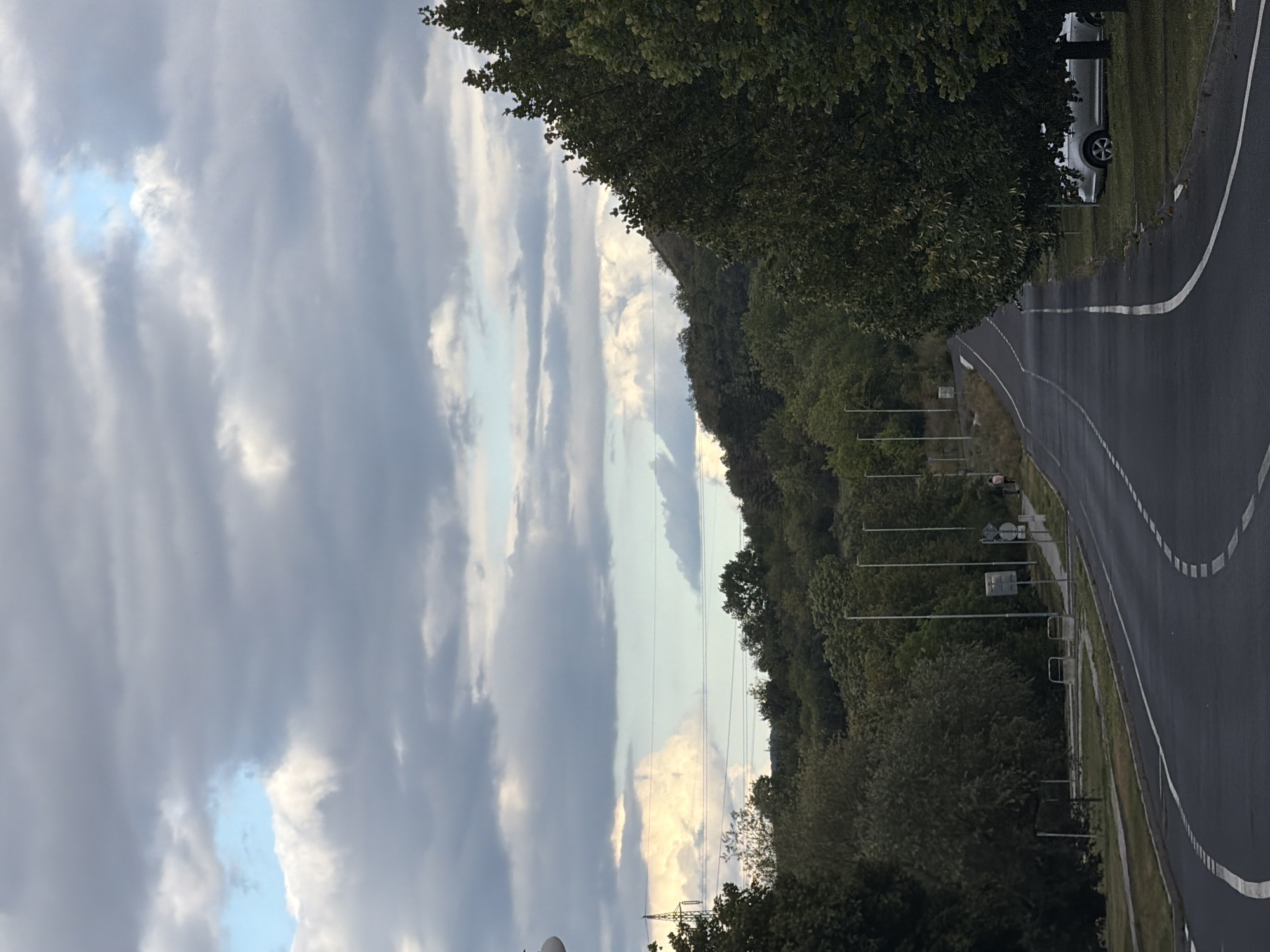 The image depicts a road in Louny, Czechia, bordered by trees and a hilly landscape under a cloudy sky. The road curves to the right, with white dashed lines marking the lanes. On the left side of the road, there is a grassy area with streetlights and signs. On the right side, several trees line the road, and a silver car is parked on the grass. In the distance, the terrain rises into a densely wooded hill. The sky is filled with various types of clouds, ranging from dark grey to lighter, more diffused formations, with a patch of blue sky visible. A person in pink is seen walking on the sidewalk on the right side of the road.