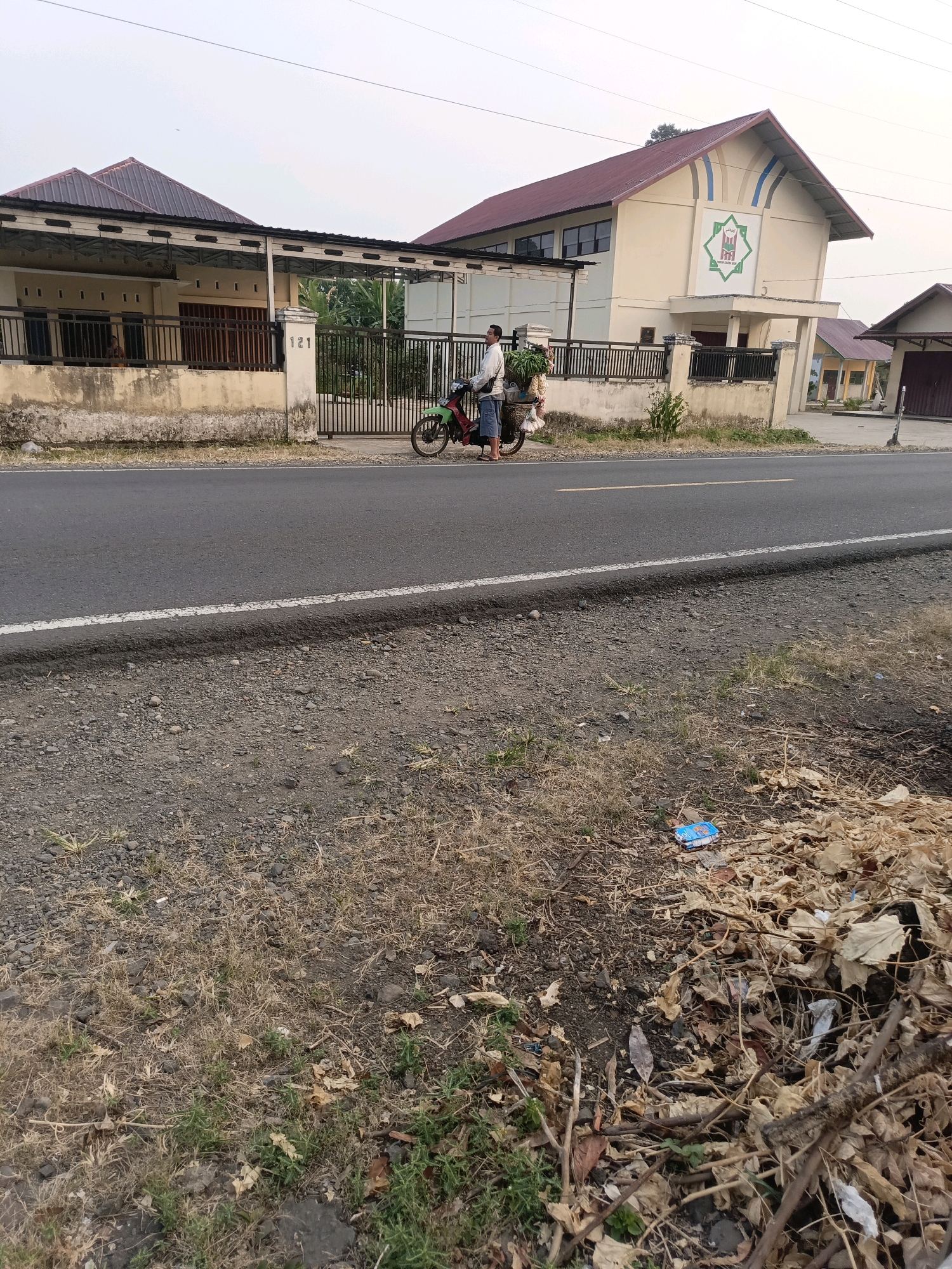 In Ulak Tanding, Indonesia, a man is seen standing next to a motorcycle, preparing to move. The motorcycle, colored green and red, is parked on the side of a paved road. The man is dressed in a white shirt and blue shorts, with a basket on the back of the motorcycle loaded with what appears to be produce. He is facing the motorcycle, his right hand near the handlebars.
To the left of the road, a building with a cream-colored facade and a dark red roof is visible. This building has a fence made of dark metal bars in front of it, and a concrete wall separates the property from the road. A sign with the number "121" is mounted on a pillar near the gate.
Further to the right, across the street, stands another building with a similar cream-colored facade and a prominent dark red roof. This building features a large emblem on its front, composed of a green star-like shape with internal detailing. A fence similar to the one on the left separates this property from the road.
The environment surrounding the buildings and road appears to be a mix of dry vegetation, including grass and fallen leaves, with some scattered debris on the roadside. The sky is overcast, suggesting a cloudy or hazy day.
