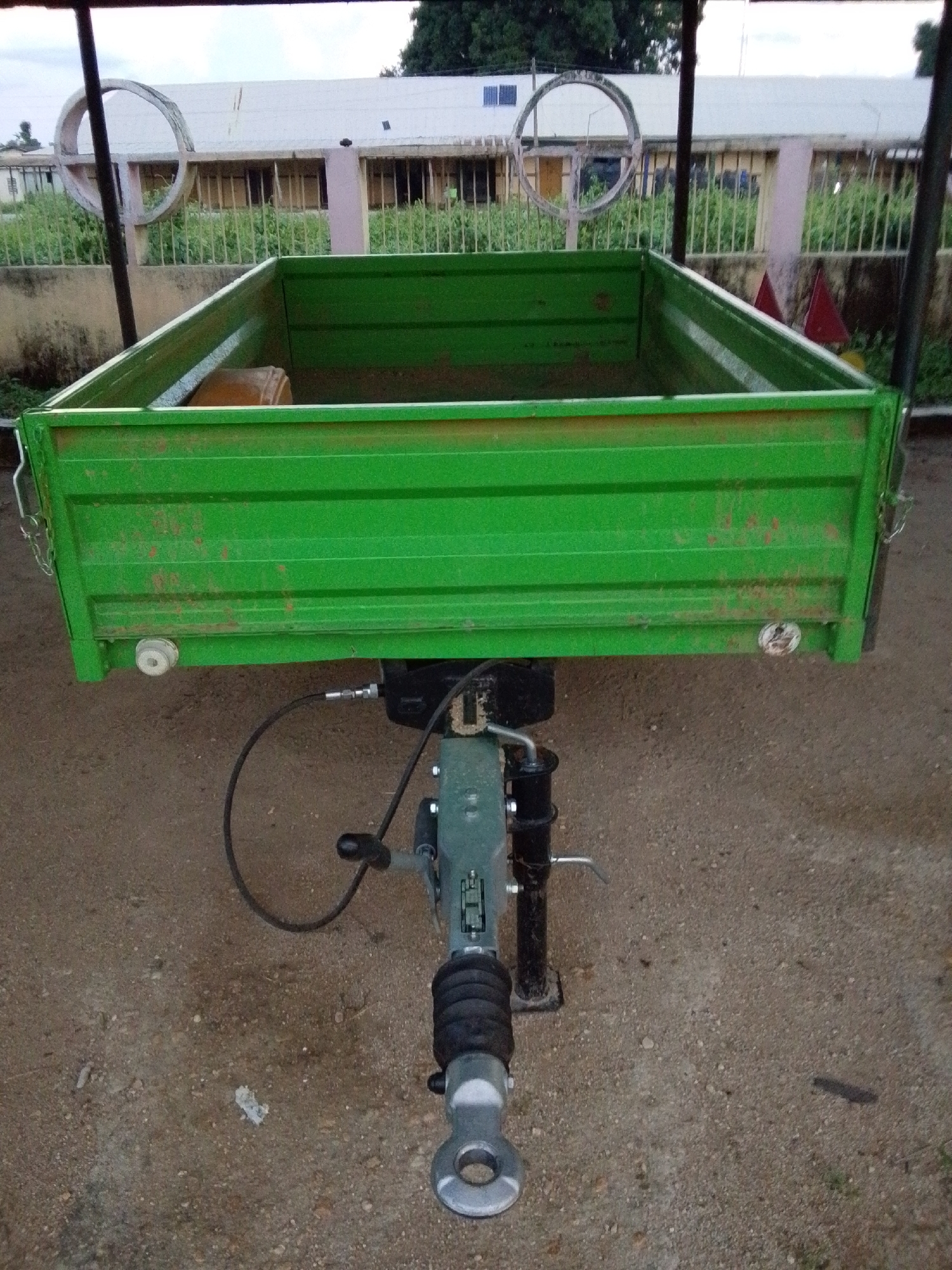 The image shows a green, metal-sided trailer with a trailer hitch mechanism in the foreground. The trailer bed is empty except for a light brown, cylindrical object. The trailer is parked on a gravel or dirt surface. In the background, a white building with barred windows and an overgrown fence with green foliage is visible. A large, circular, metal structure, possibly a decorative element or part of a larger installation, is present near the building. The sky is overcast. The scene appears to be an outdoor area, possibly a yard or storage space, in Serti, Nigeria.
