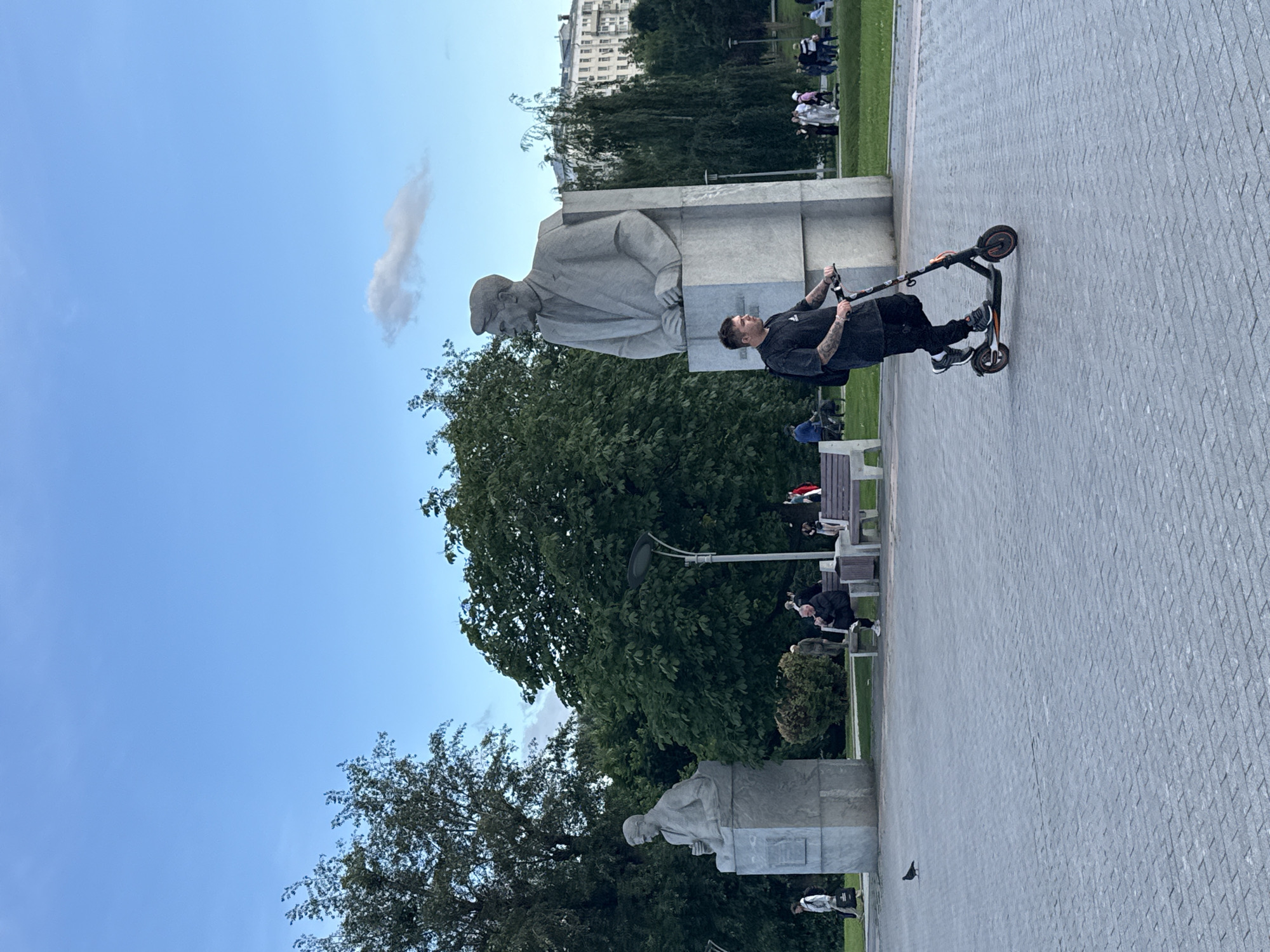 This media file depicts an outdoor scene in Moscow, Russia, likely during the daytime with clear skies and moderate weather. The primary subjects are people engaged in various activities within a park-like setting.
In the foreground, a person is seen riding a scooter. This individual is dressed in dark clothing and is focused on navigating the paved pathway. The scooter itself is a modern, electric model.
Dominating the midground are large, grey stone statues. One statue appears to be a bust of a man in a cap, set against a substantial monument. Another, smaller statue is visible further in the background, also appearing to be of a person. These statues suggest the location is a place of historical or cultural significance, possibly a memorial park.
Around the statues, several other individuals are present. Some are seated on park benches, while others are standing or walking. Many appear to be casually observing the surroundings or interacting with each other. The presence of benches and manicured green spaces indicates a public park or plaza designed for recreation and relaxation.
In the background, a tall, multi-story building with numerous windows can be seen, characteristic of urban architecture in Moscow. The overall impression is that of a bustling public space where people are enjoying a pleasant day outdoors, with a mix of modern elements like the scooter and historical markers like the statues. No visible text is present in the image.