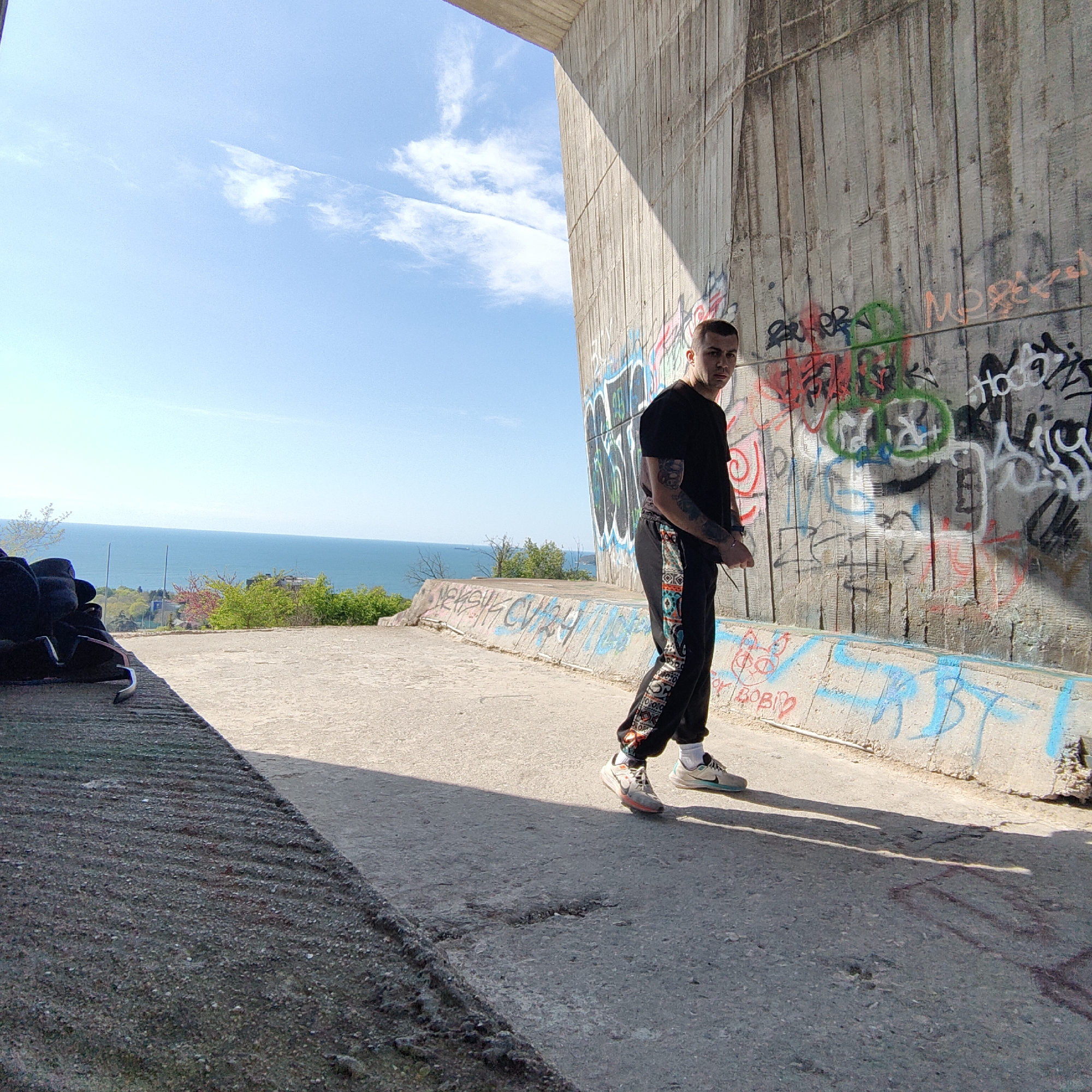 A medium shot features a young man in Varna, Bulgaria. He has short, dark hair and tattoos on his arms and wears a black t-shirt and patterned black pants. He stands on a concrete surface next to a graffiti-covered concrete wall. The background shows a blue sky with a few clouds and the Black Sea. The photo captures the urban environment of Varna, highlighting its architecture and street art.