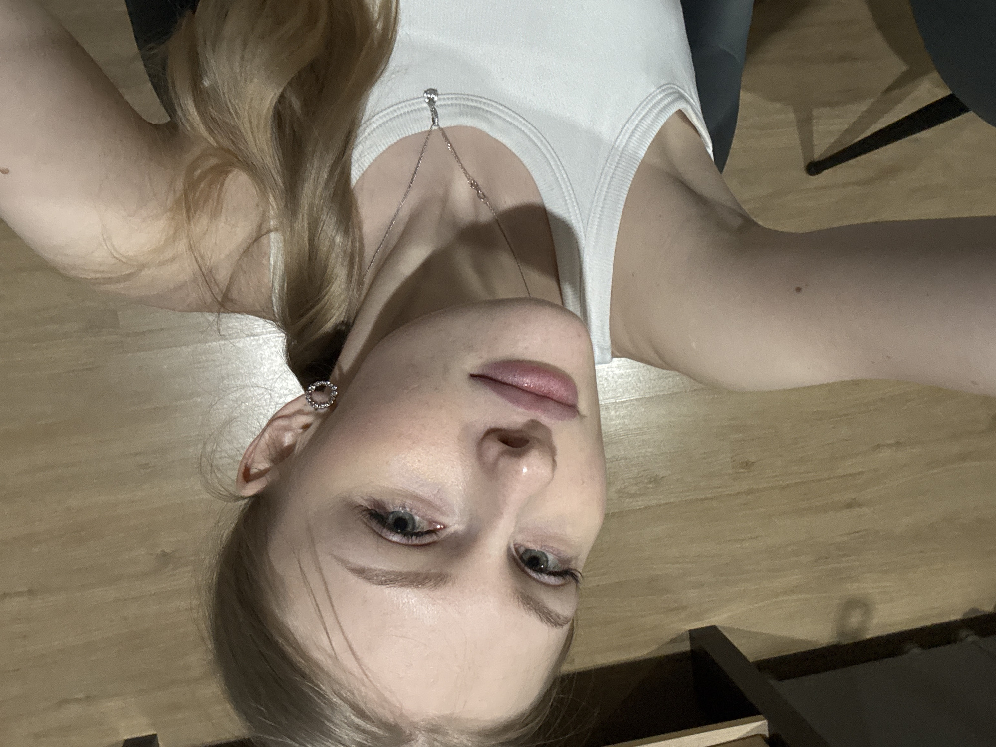 A close-up, vertically oriented selfie captures a young woman lying on her back, looking directly up at the camera. Her head is positioned at the bottom of the frame, with her upper body and arms extending towards the top. She has long, wavy, light blonde or light brown hair draped around her, and fair skin. Her eyes are light-colored, appearing to be greenish-grey or blue, accented with eyeliner, and her eyebrows are well-defined. Her lips are slightly parted and appear to have a light gloss.
She is wearing a white ribbed tank top. A delicate silver-colored chain necklace with a small, reflective pendant rests against her chest. A circular, silver-toned earring is visible in her left ear. Her right arm is visible on the left side of the frame, showing a small mole on the bicep area.
The background consists of a light-toned wooden floor with visible plank patterns, indicating an indoor setting. A dark, angular object, likely a furniture leg, is partially visible in the upper right corner, casting a shadow on the floor. The lighting appears soft and even, suggesting indoor illumination. The woman's expression is neutral and direct. There is no visible text in the image.