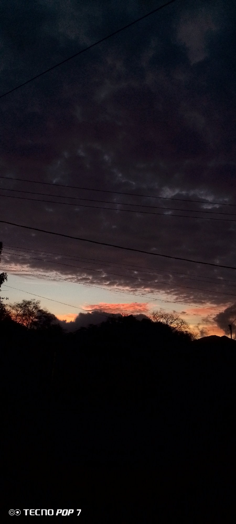The image shows a landscape at dusk or dawn in Altagracia, Venezuela. The sky is filled with dark, heavy clouds, except for a horizontal strip of orange and pink hues just above the horizon. Silhouetted trees and hills occupy the lower portion of the image. Power lines run across the sky. The bottom of the image has the text "TECNOP POP 7".