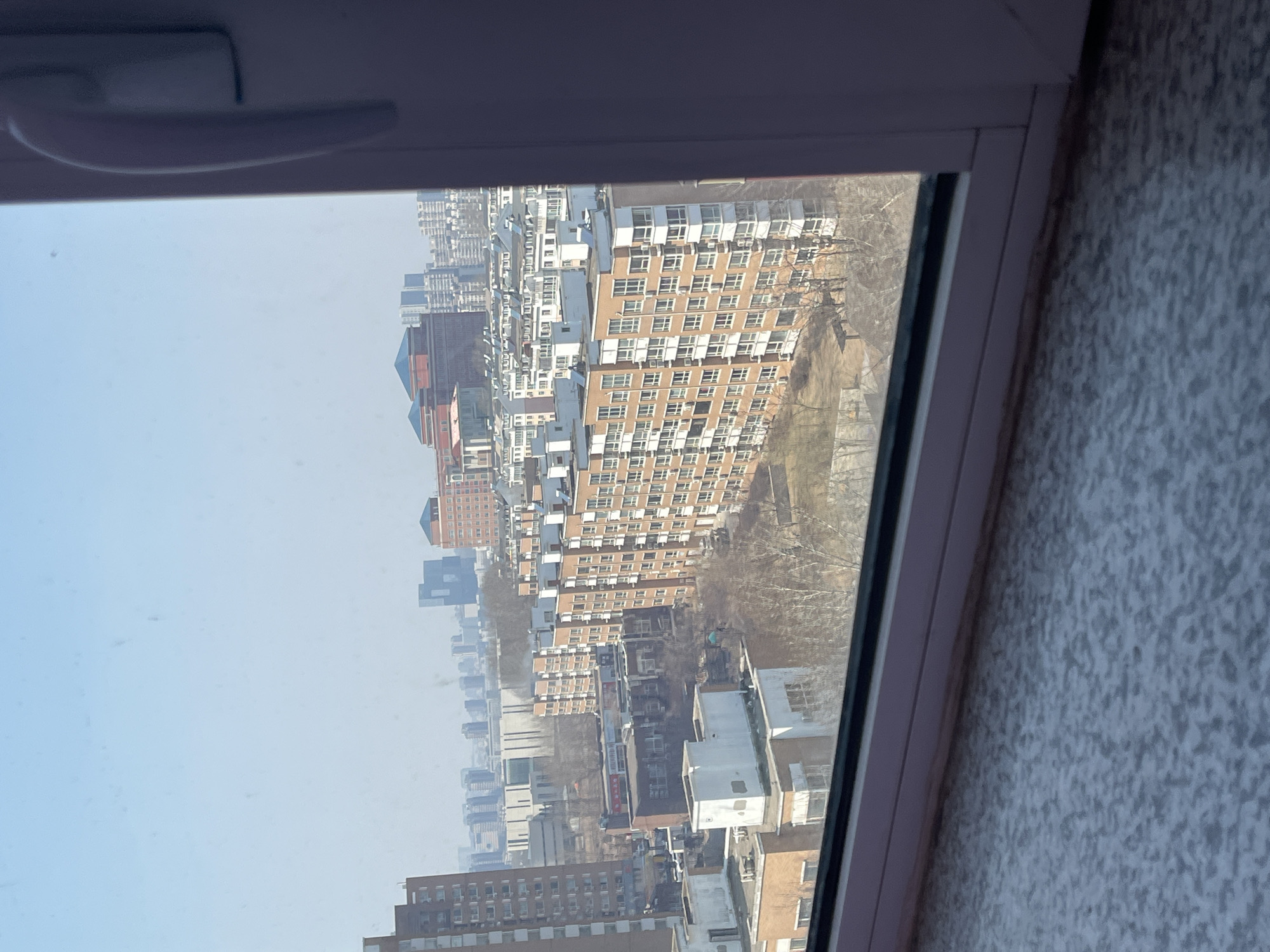 The image displays a daytime urban panorama of Changchun City, China, viewed through a window. The immediate foreground features a speckled gray-blue surface, likely a windowsill, and a partial white window frame with a handle on the right side. The scene extends under a clear, pale blue, somewhat hazy sky.
In the midground, a dense arrangement of multi-story buildings is visible. A prominent light brown residential building with white balconies, approximately 8-10 stories high, occupies the right-center of the frame. To its left, a taller, darker brown apartment block is partially visible. The ground level areas between buildings contain bare deciduous trees and dry, patchy earth, suggesting a late winter or early spring season. Some rooftops display what appear to be solar water heaters.
The background reveals a further expanse of the city, characterized by numerous high-rise buildings and distant skyscrapers that are partially obscured by atmospheric haze. Notable structures include a large, red-hued building with distinctive blue, dome-shaped roofs in the upper-right background, and a modern, dark-colored, irregularly shaped building to its left. No individuals, specific activities, or interactions are discernible within the scene.