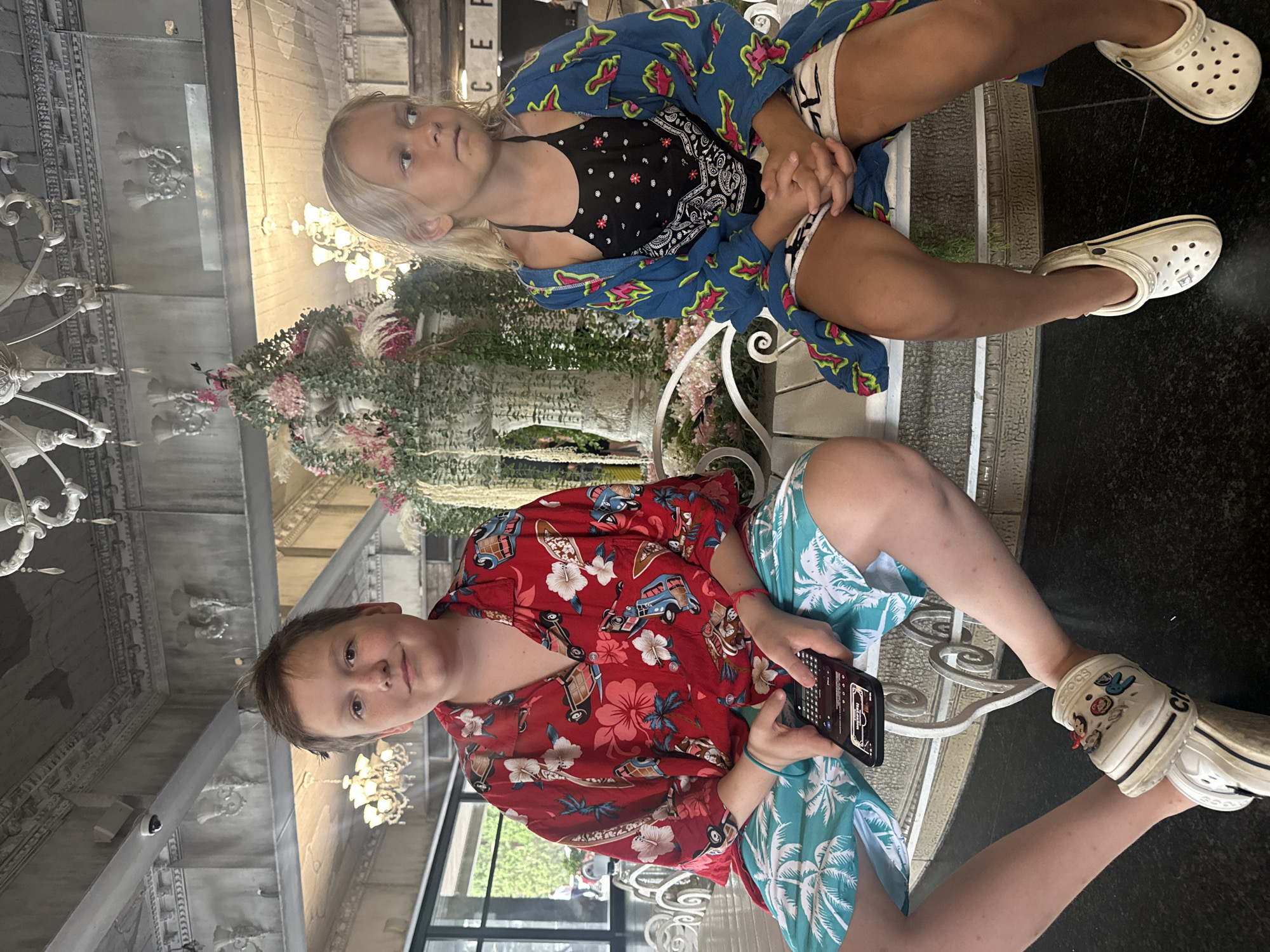 Two children are sitting on a bench in what appears to be a hotel lobby. The child on the left is a boy wearing a red Hawaiian shirt with a floral and car print, and blue and white palm tree patterned shorts. He is holding a smartphone. The child on the right is a girl wearing a blue patterned robe over a black bandana-print tank top and white shorts. Both children are wearing white Crocs. Behind them is a large floral fountain and ornate ceiling decorations. The background suggests an indoor setting, possibly a resort or hotel, in Yalta, Ukraine.