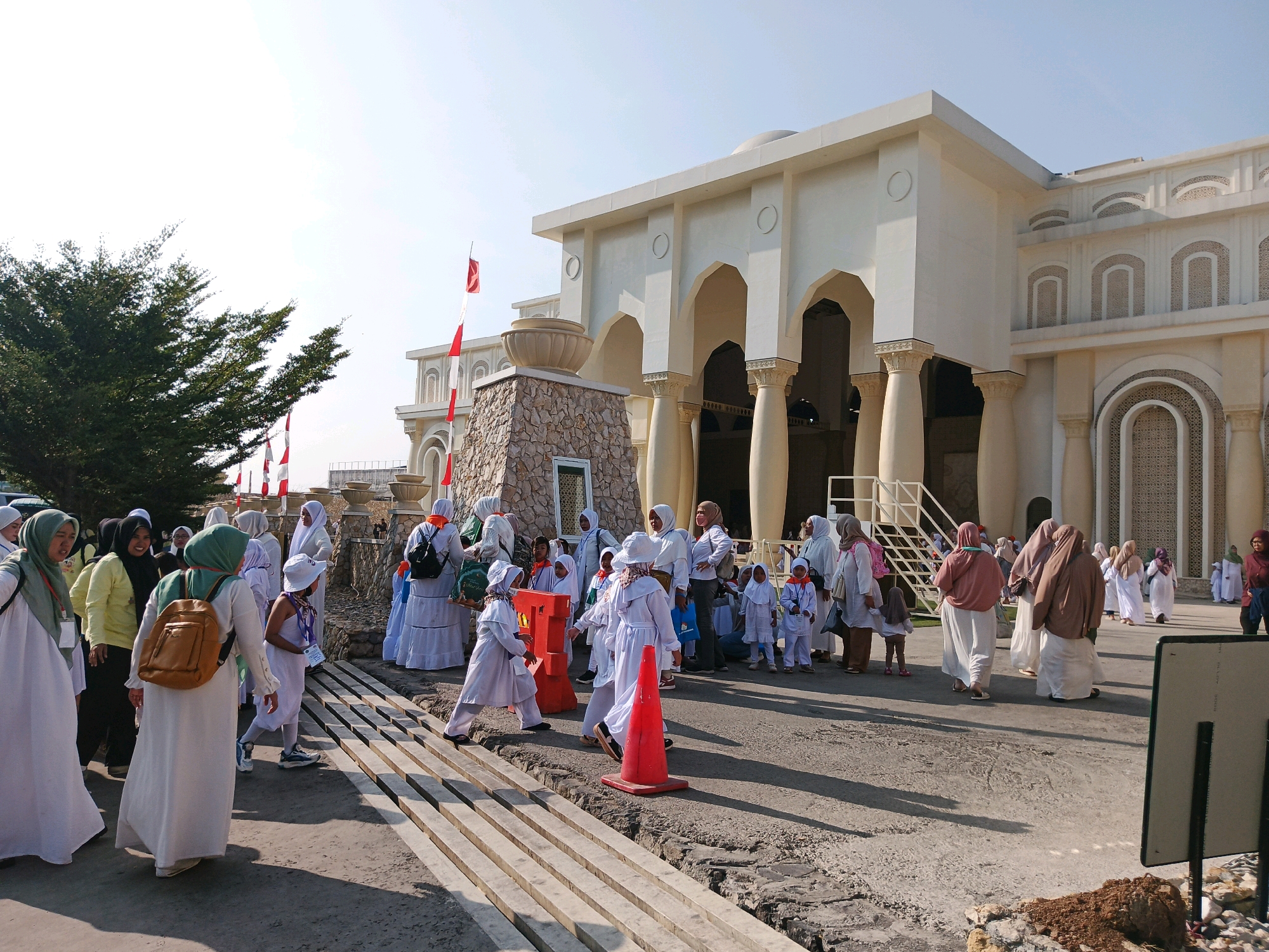 The image depicts a group of people, many wearing white and light-colored Islamic attire, gathered in front of a large, modern building with classical architectural elements, possibly a mosque or a government building. The building is cream-colored with arched doorways and colonnades. Several Indonesian flags, red and white, are visible, one prominently on a flagpole to the left of the building. The ground is paved, and there are wooden planks forming a walkway or steps. In the foreground, a few individuals are walking, and some are observing the surroundings. A bright orange traffic cone is placed on the ground. The overall atmosphere suggests an outdoor gathering, possibly a religious event or a public ceremony, in a sunny climate. The presence of the Indonesian flags strongly suggests the location is in Indonesia, and the architecture, while having classical influences, is contemporary. Given the visual cues of the building's design, the flags, and the attire of the people, this scene could be located within a prominent public or religious site in Bandung City, Indonesia.