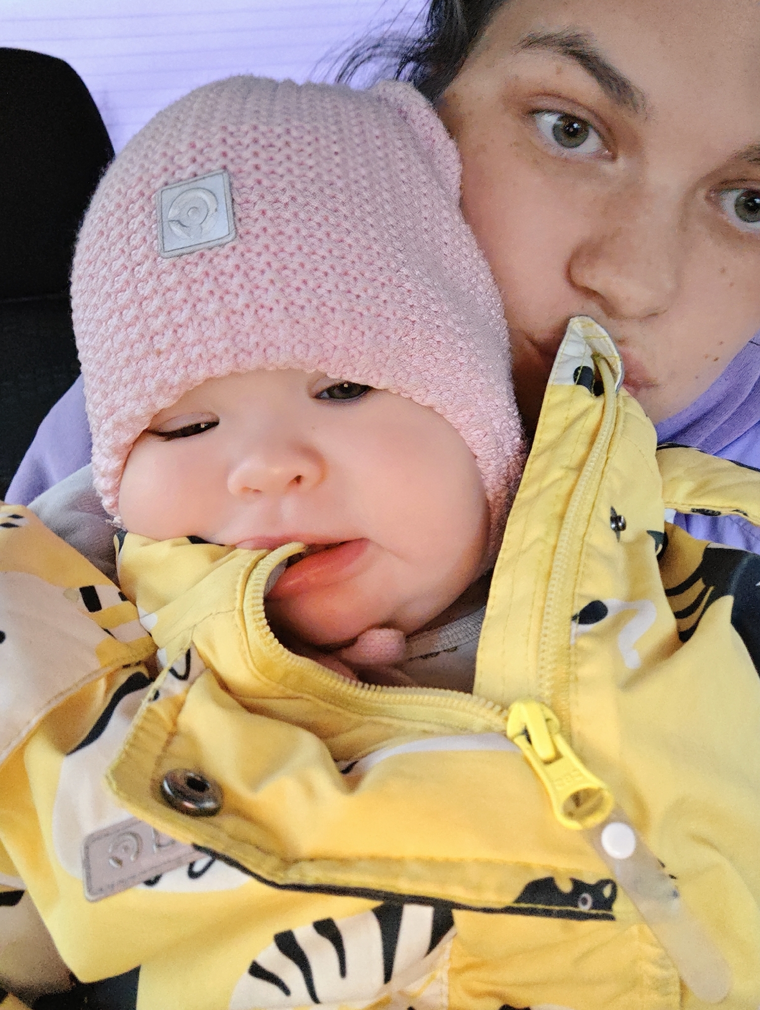 A close-up shot depicts a baby in a pink knit hat, wearing a yellow jacket. The baby is being held by an adult, who is kissing the baby's jacket. The baby is looking at the camera. The location is not discernible from the image.
