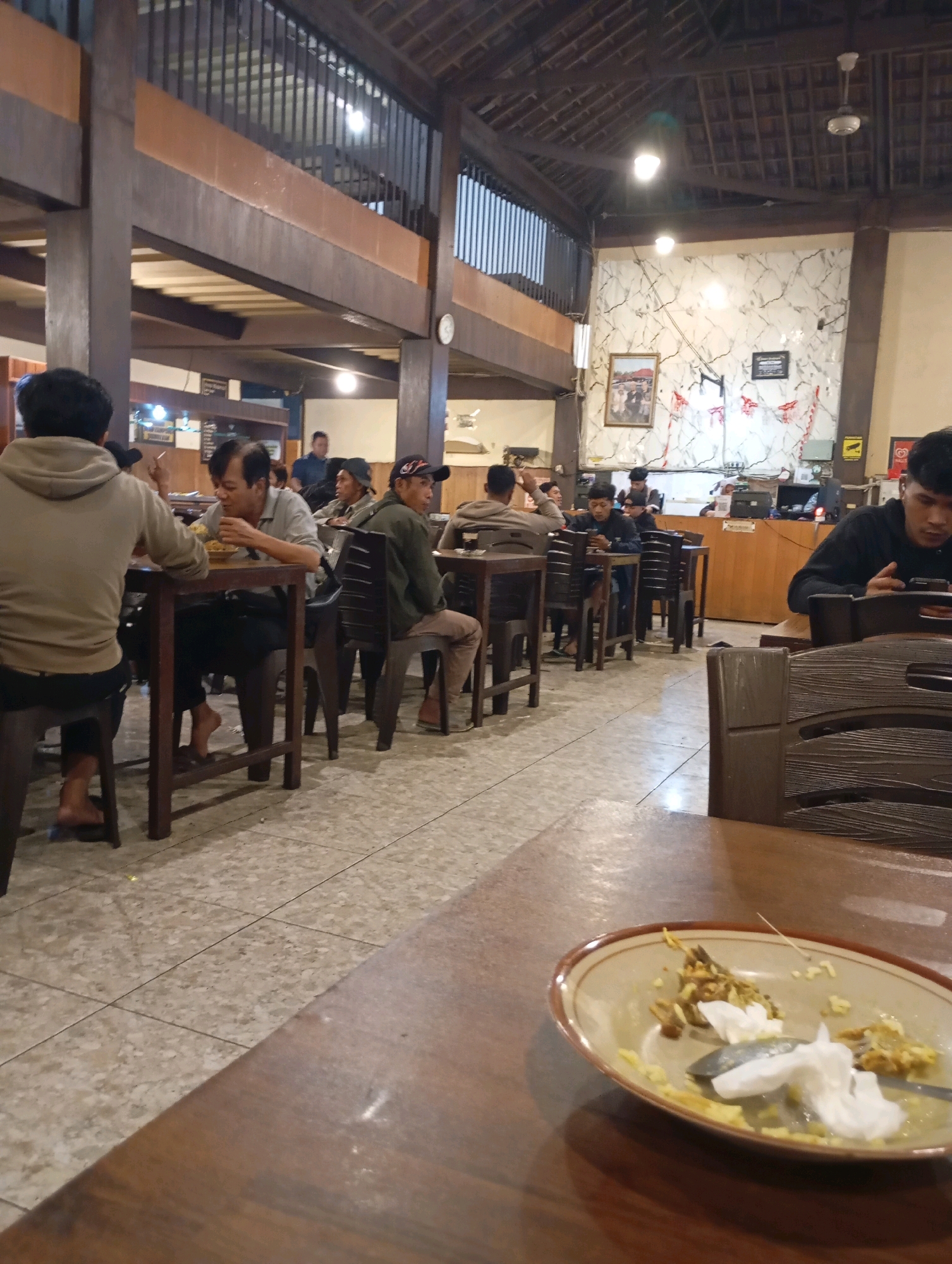 The image depicts the interior of a restaurant, possibly in Purworejo, Indonesia, with a rustic and traditional architectural style. The ceiling is made of wood with exposed beams and a tiled roof structure. The walls are a mix of plain white and dark wood paneling, with some decorative elements like framed pictures and flags. The floor is tiled.
Several patrons are seated at wooden tables, engaged in eating or observing their surroundings. In the foreground, a partially eaten meal is visible on a plate on a wooden table. In the midground, several men are seated at tables, some eating, others looking at their phones. One man in the foreground is wearing a light brown hoodie, another in a light grey shirt is eating. To the right, a man in a dark hoodie is looking at his phone. The overall atmosphere appears to be casual and relaxed.