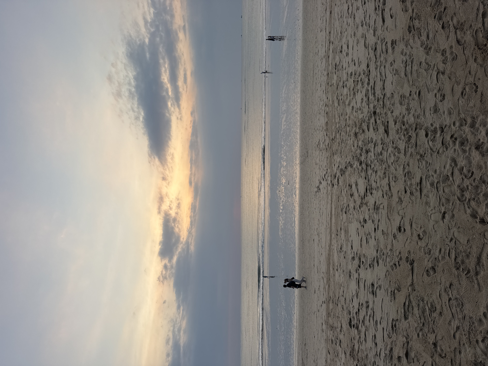 A serene beach scene unfolds at sunset in Kuta Selatan, Indonesia. The sky above is a canvas of soft pastel hues, with warm oranges and yellows bleeding into pale blues and grays, punctuated by wispy clouds. The ocean stretches out to the horizon, its surface a mirror reflecting the muted colors of the sky. Gentle waves lap the shore, creating a thin, wet sheen on the sand.
On the wet sand near the water's edge, two figures walk together, their silhouettes dark against the brightening wet sand. Further out, a solitary figure stands near the water. On the dry sand in the foreground, numerous footprints are imprinted, suggesting recent activity. The beach itself is composed of fine, light-colored sand. The overall impression is one of tranquility and the peaceful end of a day at the beach.
