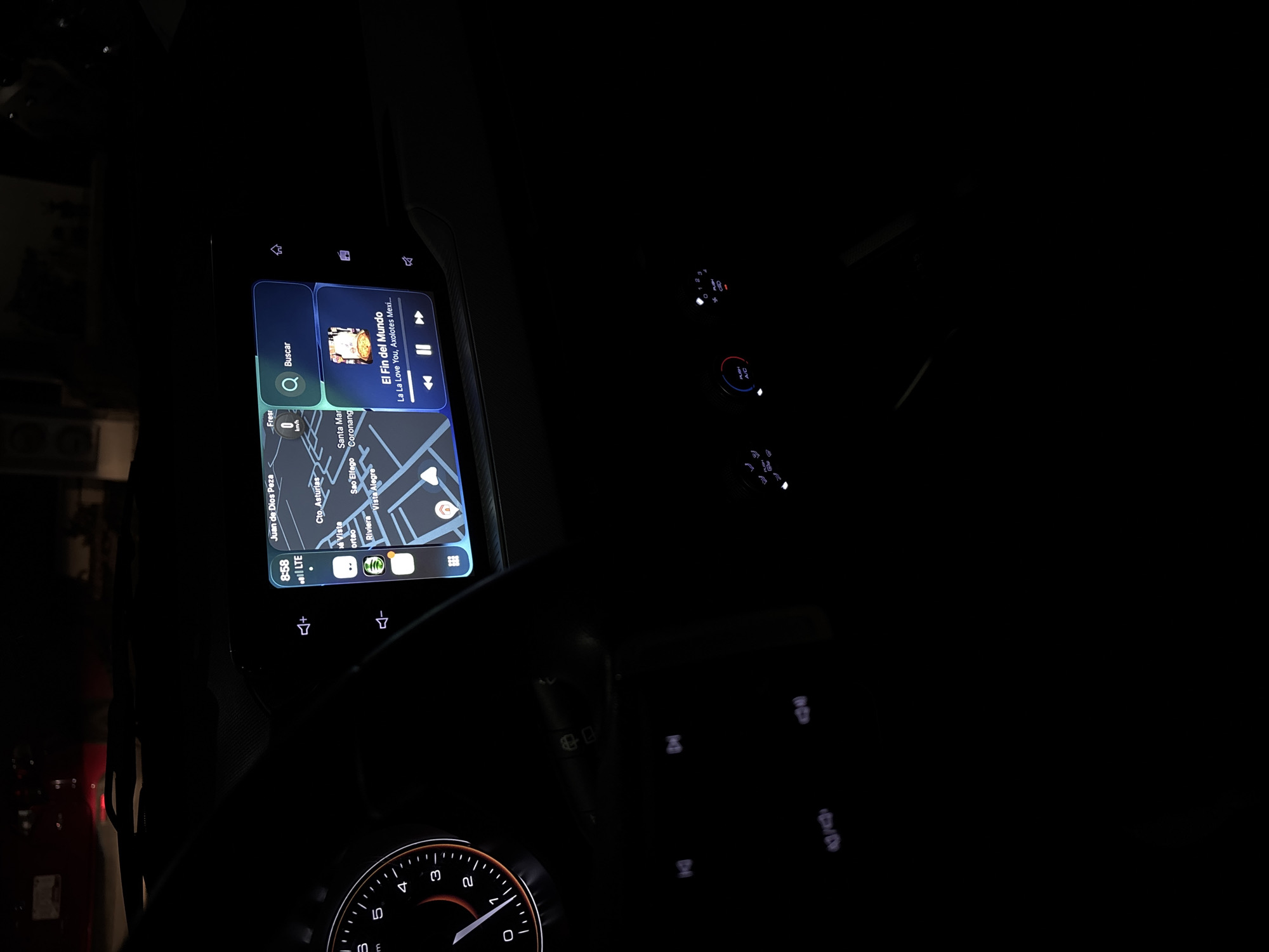 Here is a concise, forensic, matter-of-fact description of the scene:
The image shows the interior of a car at night. The dashboard screen is illuminated, displaying a map showing the location of Juan de Dios Peza, Cto. Asturias, Santa Mar, ortao, Riviera, Sao Elfego, Coronang and Vista Alegre in a location. The time displayed on the screen is 8:58, and the car is connected to LTE. The car is in park at 0 km/h. The dashboard screen is also displaying the song "El Fin del Mundo" by La La Love You, Axolotes Mexicanos. The steering wheel and a dimly lit speedometer are visible. The controls for the car's climate control system are also illuminated.