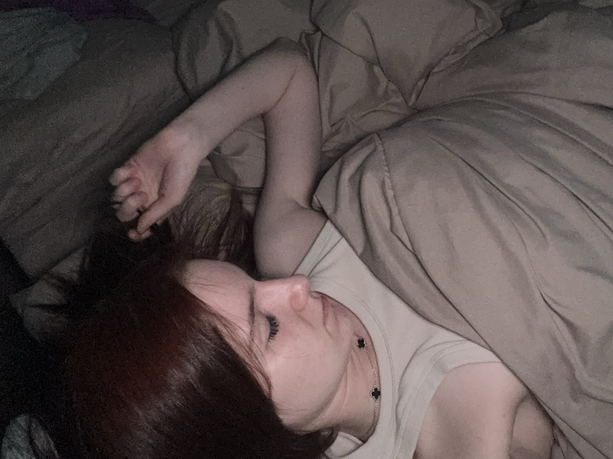 A woman is lying in a bed. She has auburn hair and is wearing a cream-colored shirt and a necklace. Her left arm is raised above her head. The bed is covered in taupe-colored sheets. The location is not discernible as Sinyavino, Russia.
