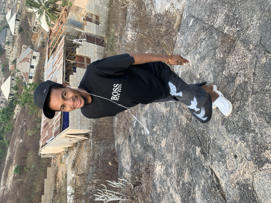 A young man in a black t-shirt with "BOSS Hugo Boss" printed on it, black cap, and patterned jeans poses on a large rock. He is smiling, wearing white shoes and earphones. In the background, there are buildings under construction and trees. The photo appears to be taken from an elevated position, possibly a hilltop or cliff.