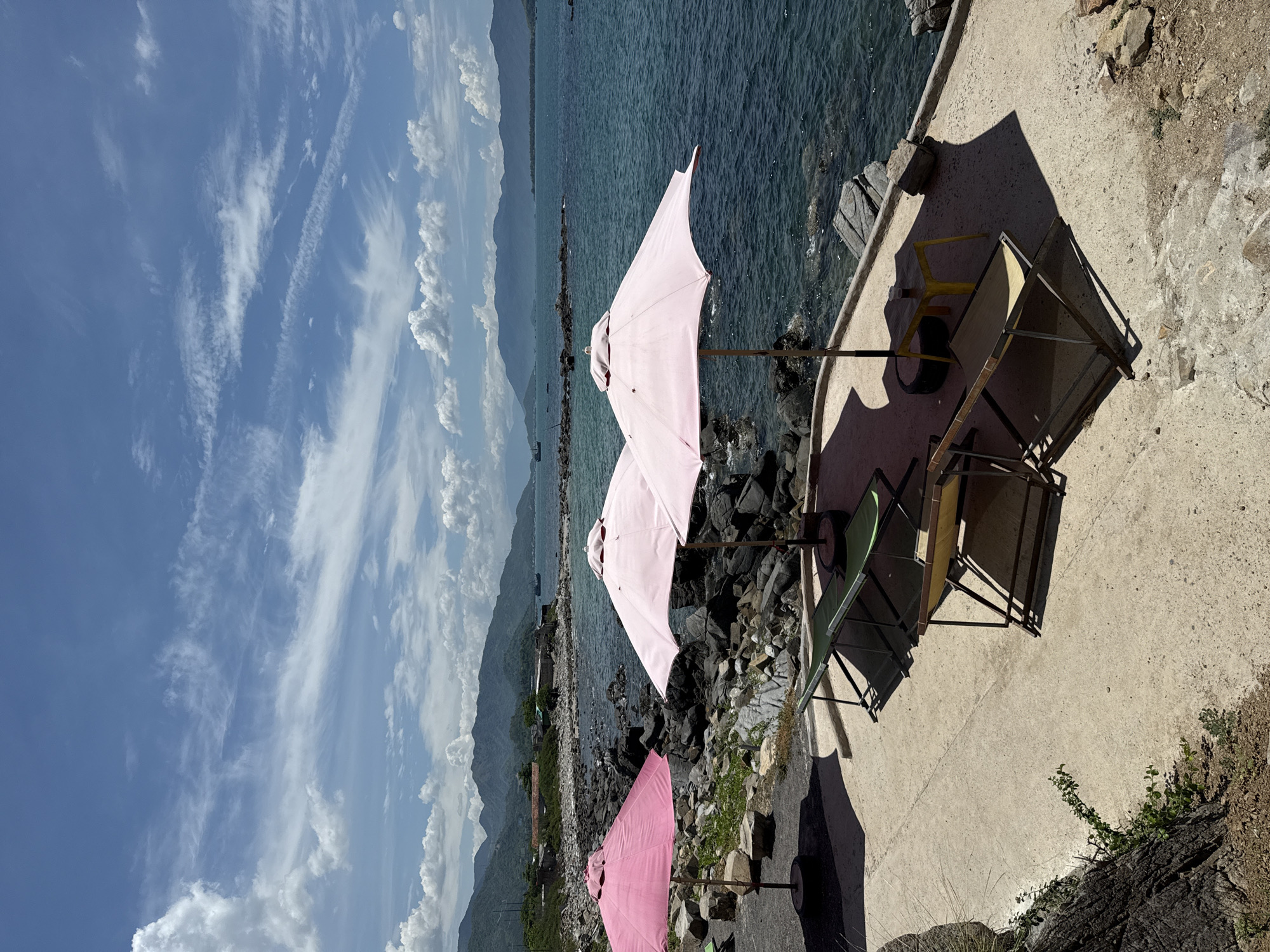 This is a photograph taken in Vĩnh Lương, Vietnam, showcasing a coastal scene with a rocky shoreline and calm turquoise water. In the foreground, a concrete pathway is lined with several pink beach umbrellas and empty lounge chairs, suggesting a recreational area. To the left, a large dark rock formation with sparse vegetation juts into the frame. The background features a hazy mountain range under a bright blue sky with scattered white clouds. The water appears relatively still, with a few small boats visible in the distance on the horizon. The overall impression is one of a serene, sunny day at a seaside location.