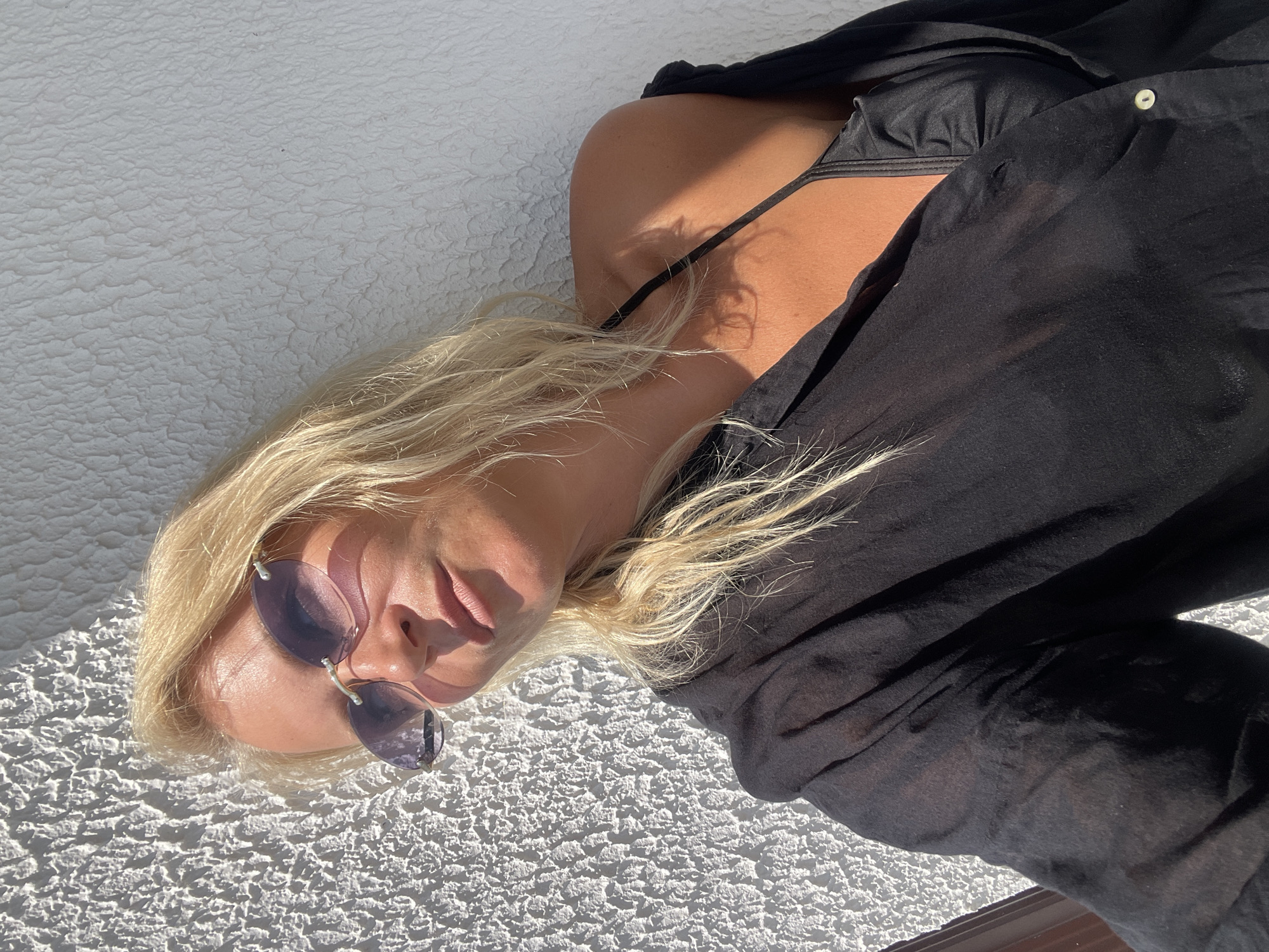 A blonde woman wearing a black unbuttoned shirt over a black bikini top and purple-tinted rimless sunglasses poses against a textured white wall. Sunlight casts a shadow across her face and illuminates her tanned skin. Her hair is loose and falls around her shoulders. The environment appears to be outdoors, possibly a balcony or patio, with a brown railing visible on the left edge of the frame. The scene is set in Giachrypsh, Abkhazia.